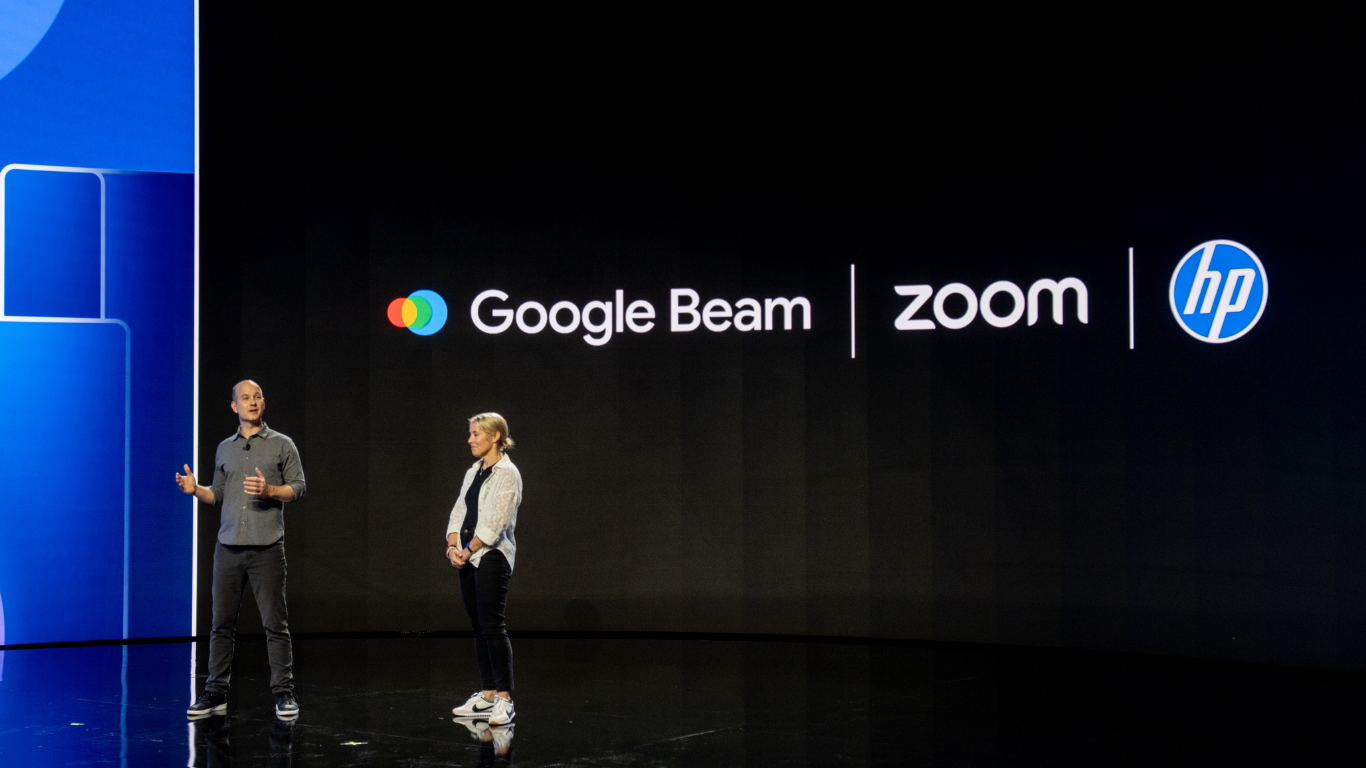 Two people on stage in front of a screen displaying the logos for Google Beam, Zoom, and HP