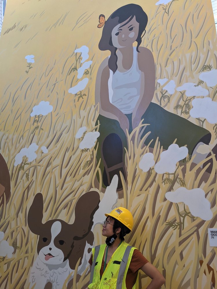 A photograph of a woman wearing a hard hat and neon vest standing in front of a mural showing yellow grasslands and a cartoon person sitting next to a cartoon dog.