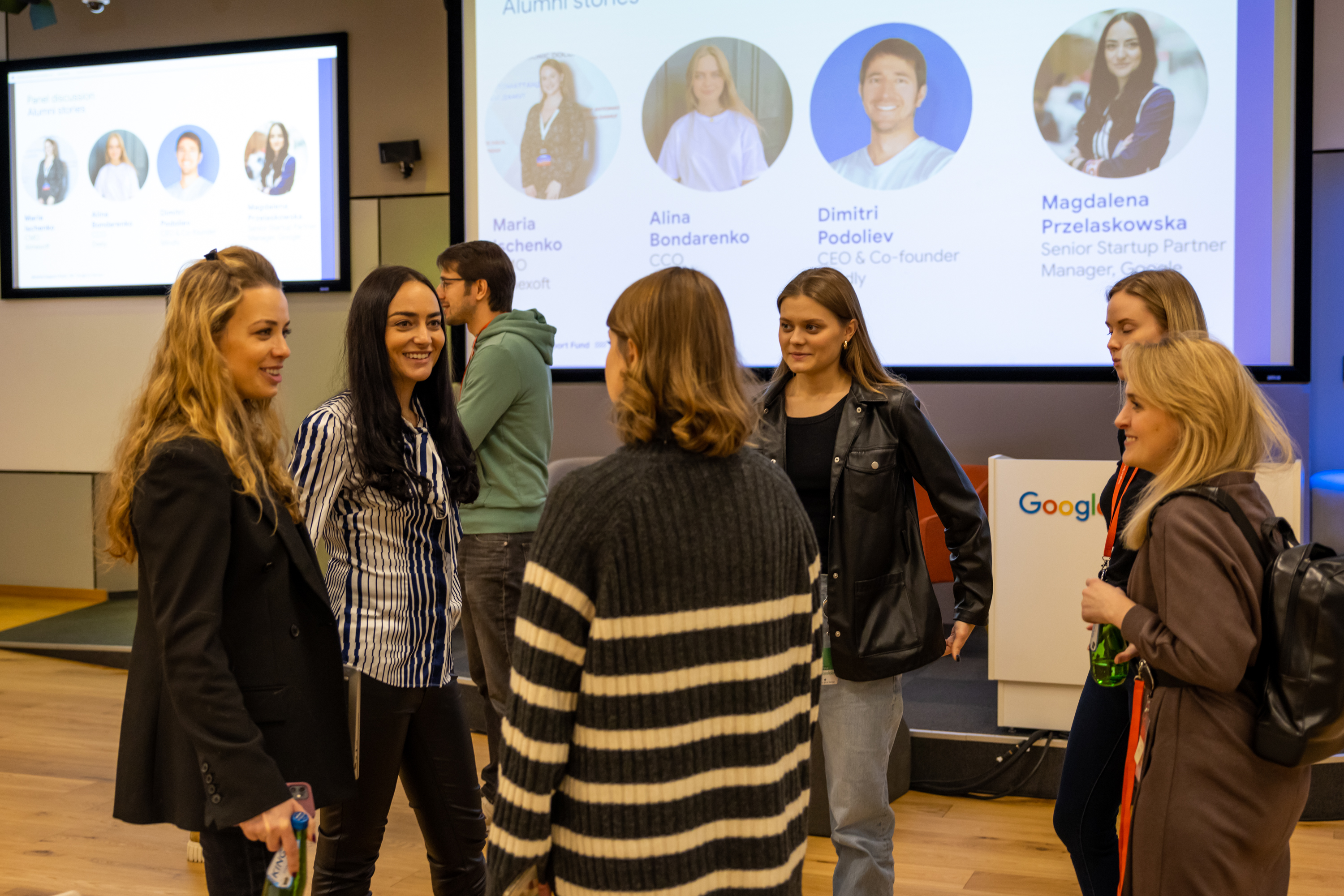 Six women are standing together in a room, smiling and talking to each other. In the background a man walks past. There is also a speaker's podium and large projector screens with four photos of people and their names and job titles.