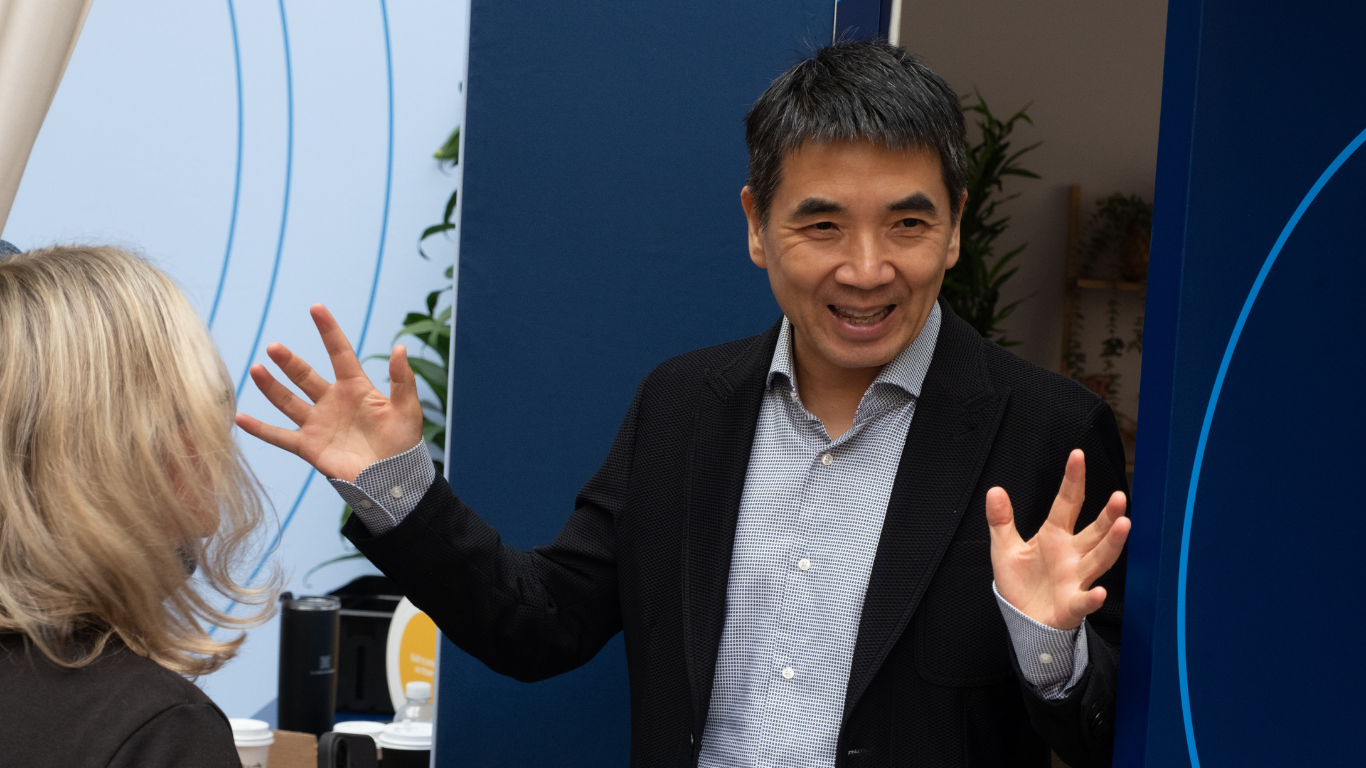 A man in a black jacket and light blue collared shirt is talking animatedly, with his hands held up, to a blonde woman.