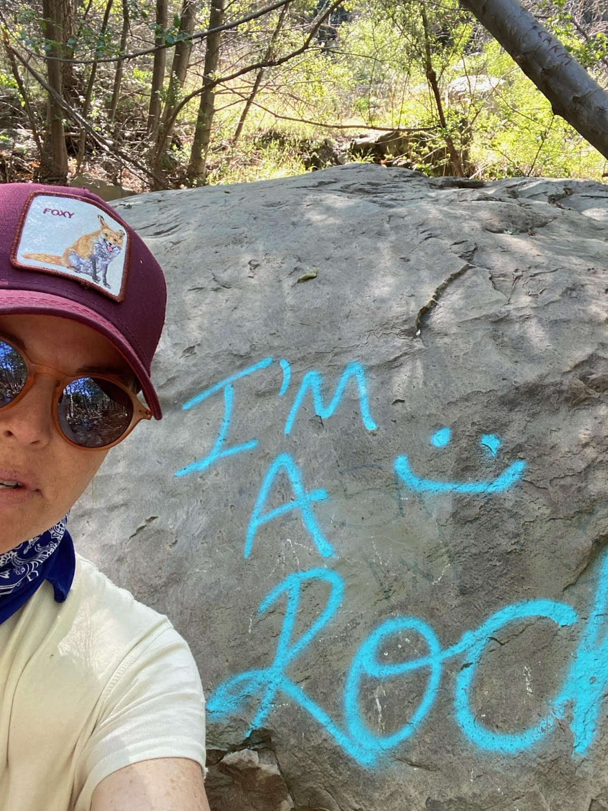 Angela posing next to a boulder that has the text "I am a Rock" painted on it in sky blue letters.