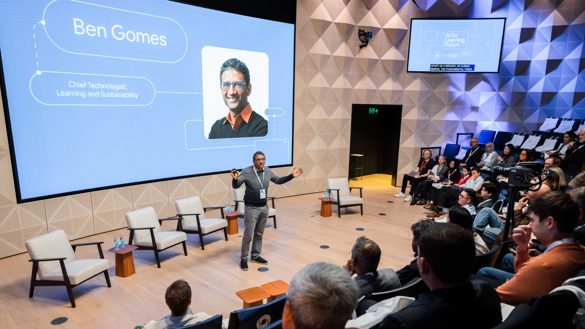 Ben Gomes presents on stage to a seated audience, with a large screen behind him displaying his name, photo, and title.