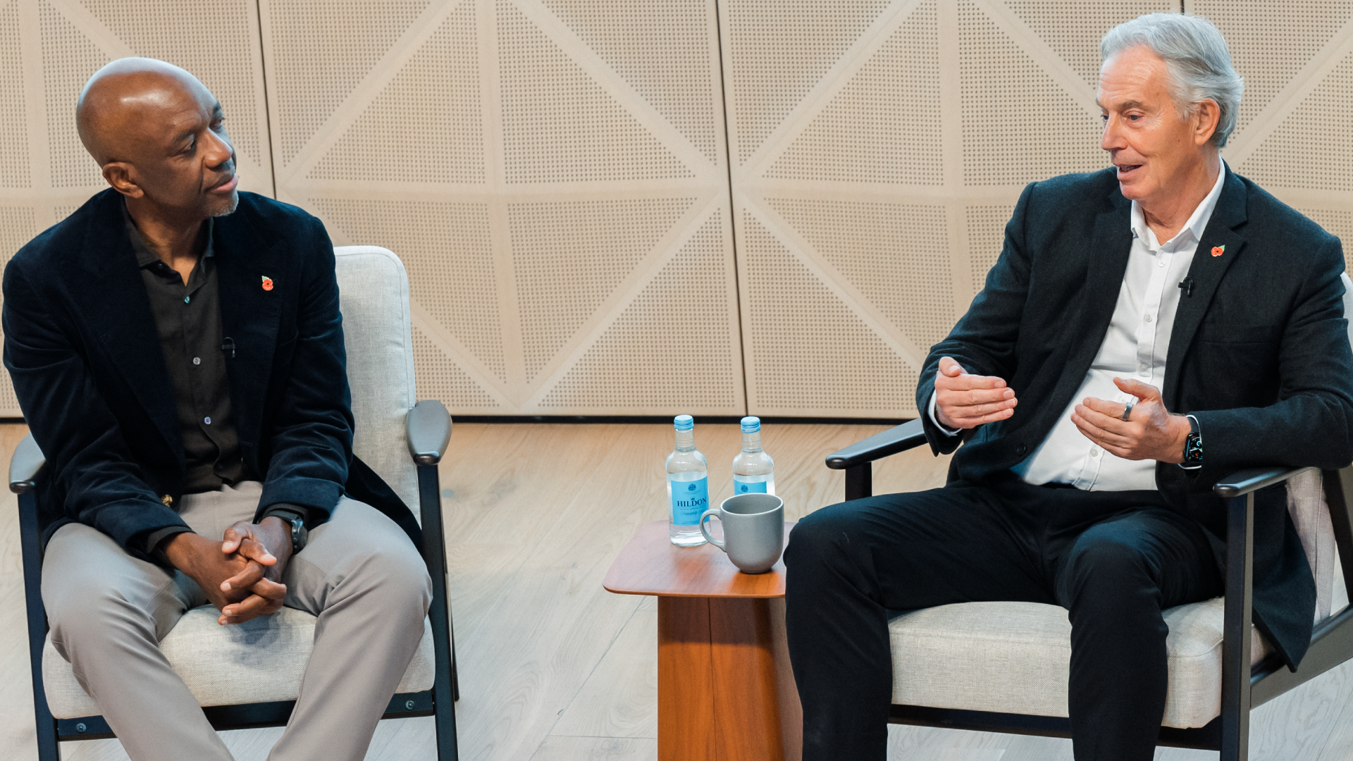 Two men seated in armchairs engage in a discussion on stage, with a small side table holding water bottles between them.