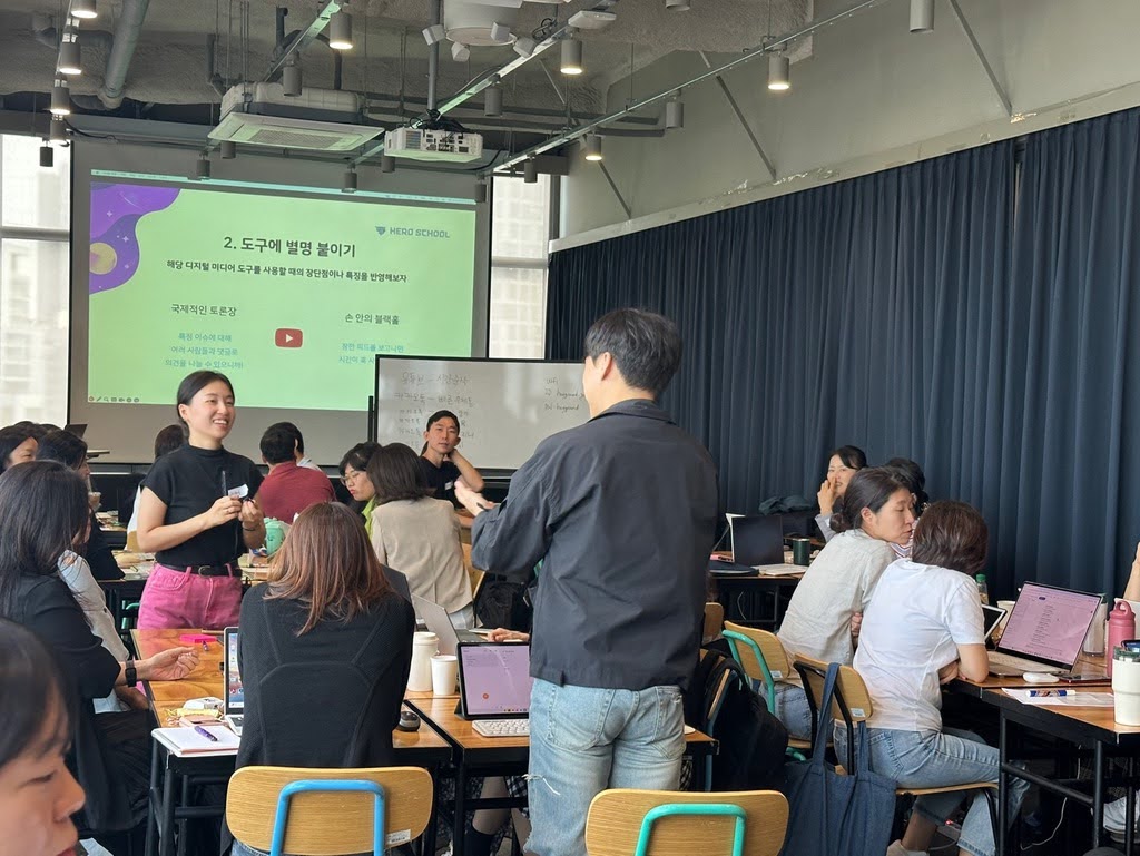A group of people are engaged in a workshop or class setting at "Hero School," where a presenter stands in front of a projection screen displaying Korean text and a YouTube icon while participants sit at tables with laptops.