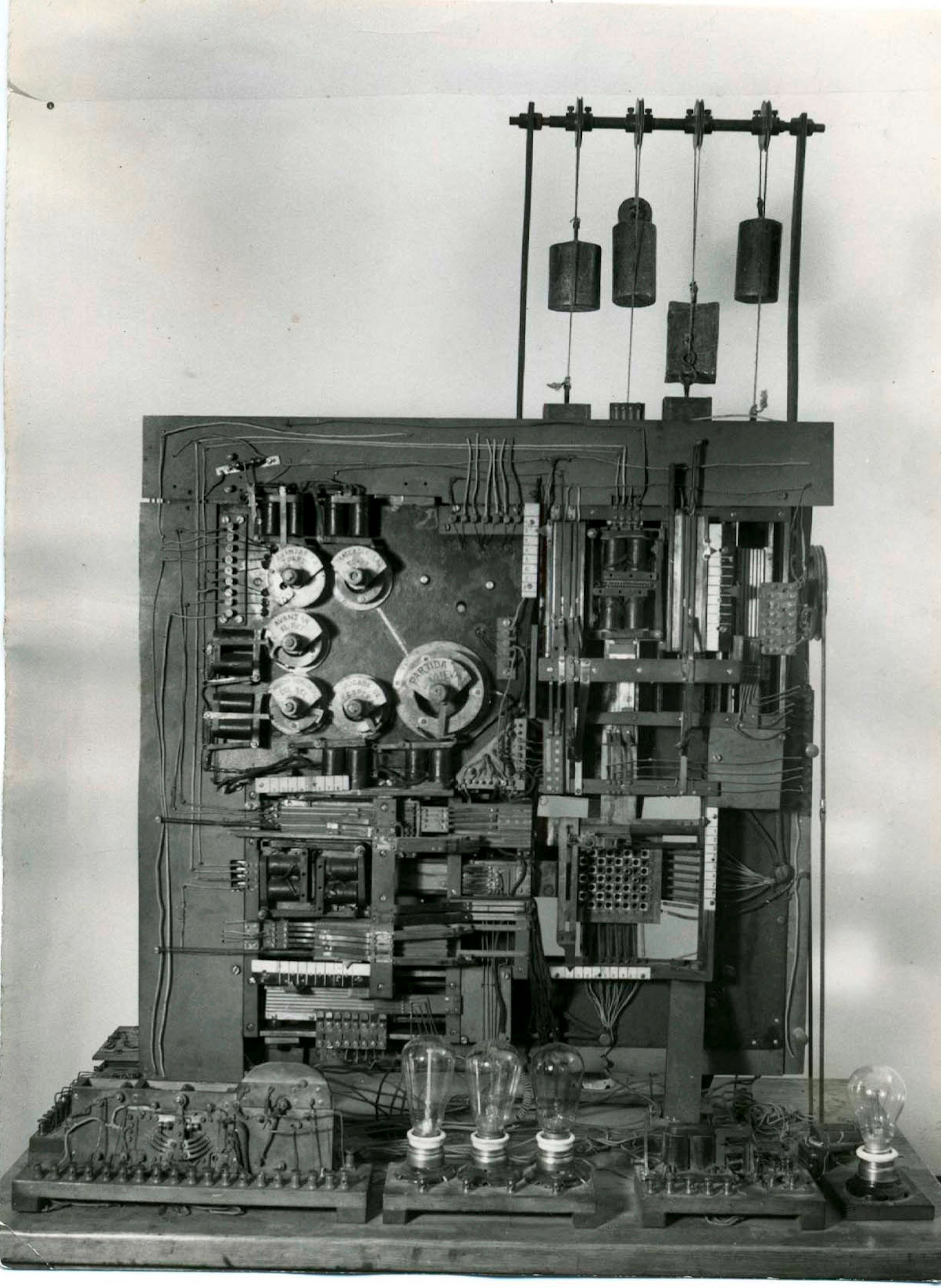 A black and white photo of a complex electronic device with wires, dials, and light bulbs