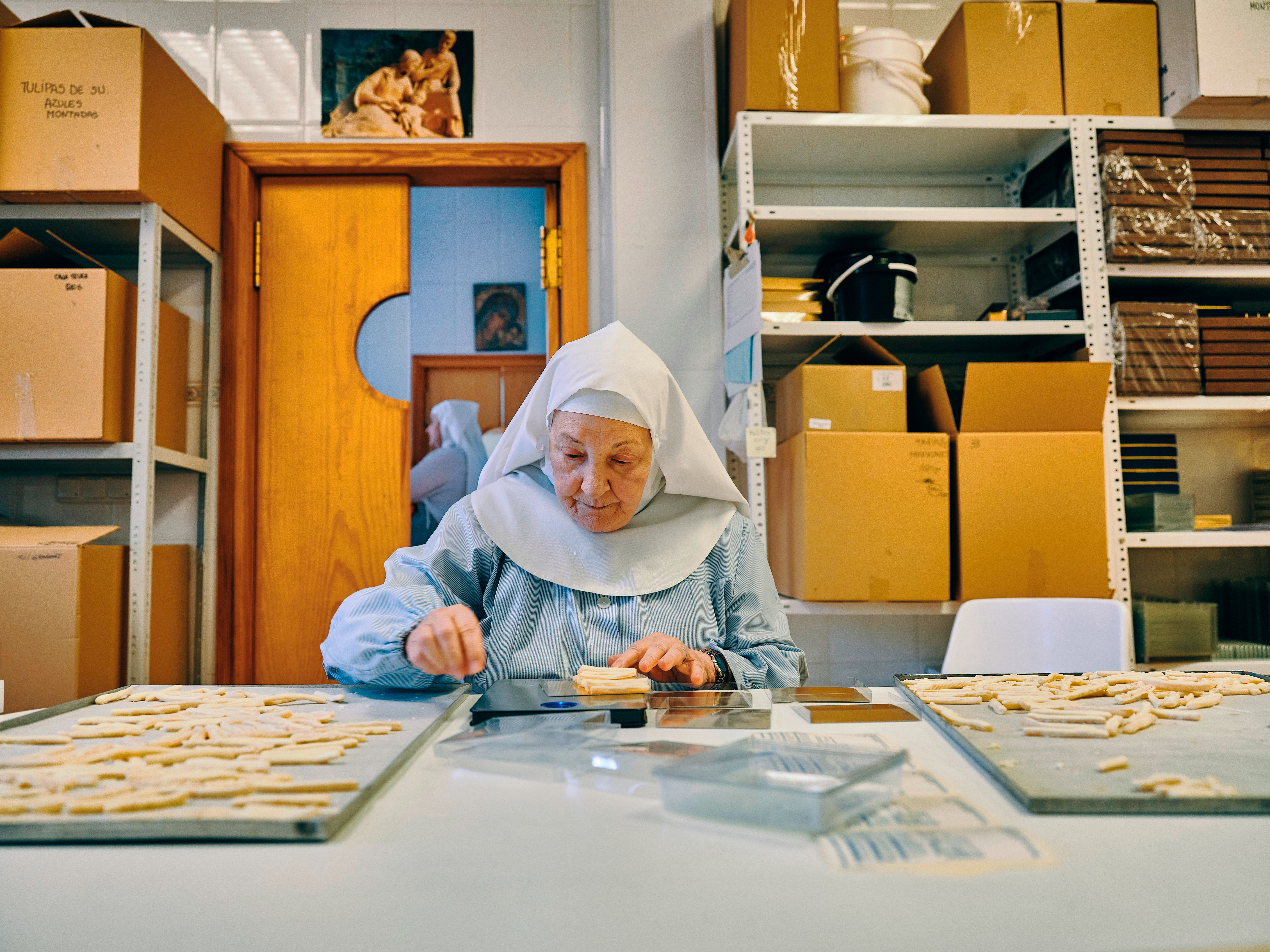 Photograph of a nun in a light blue habit, seated at a table placing small samples of chocolate on a baking tray.