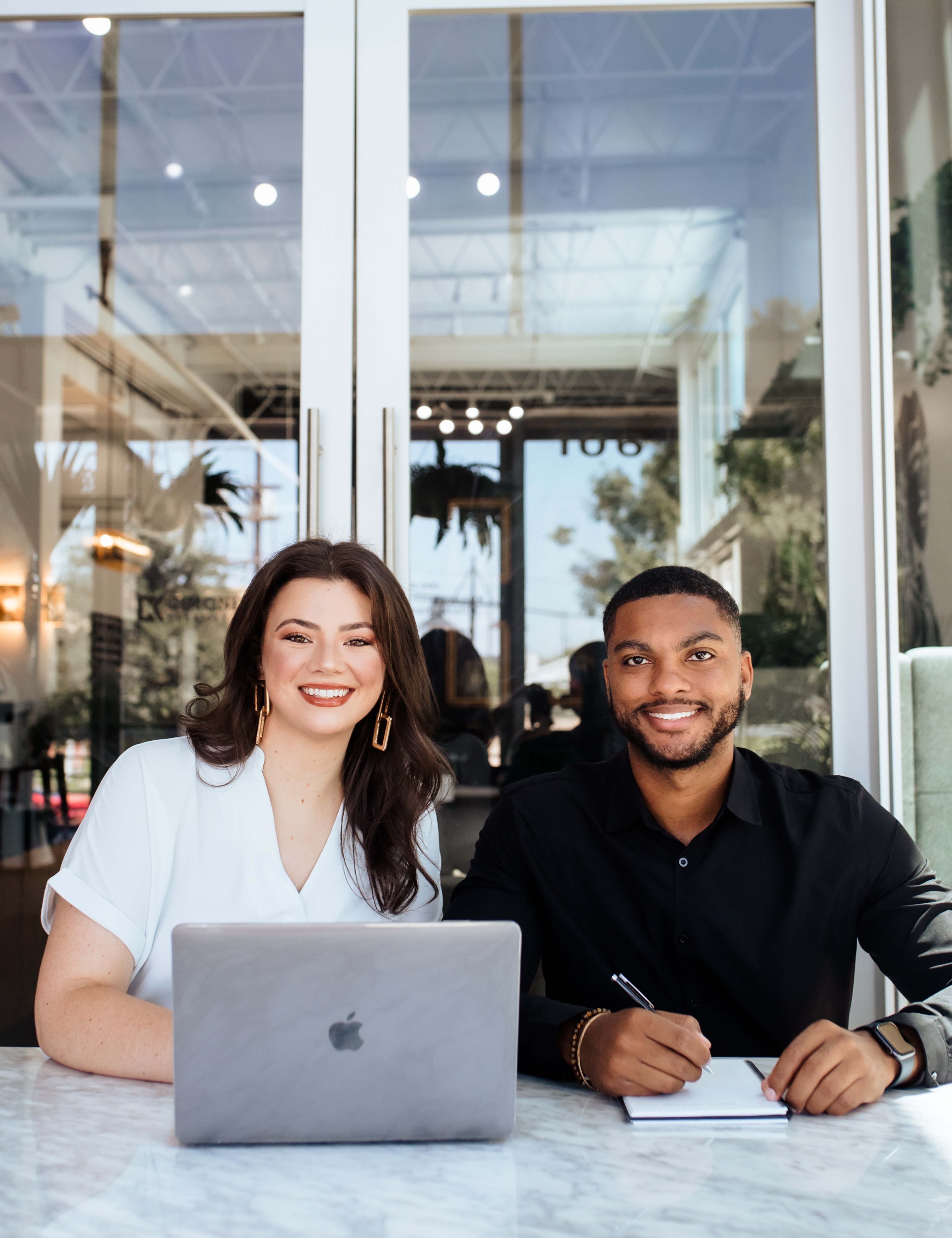 A woman in a white shirt sits next to a man in a black shirt at a white marble table. Behind them is a large glass door.