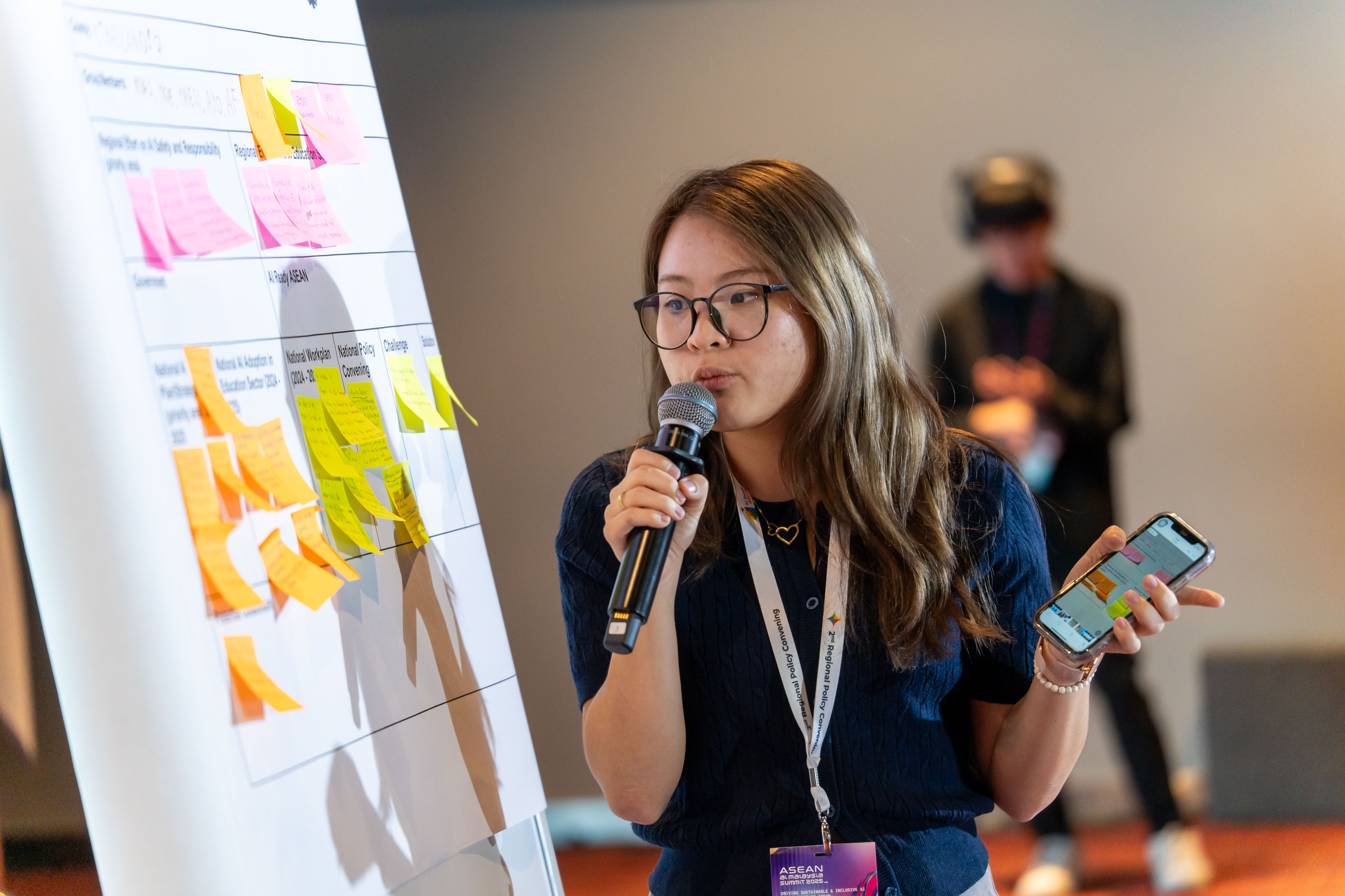 A young woman with glasses and long brown hair speaks into a microphone while holding a smartphone. She stands next to a large board covered with rows of bright pink and yellow sticky notes, suggesting a brainstorming or workshop session. Her lanyard includes an "AI Ready ASEAN" badge.