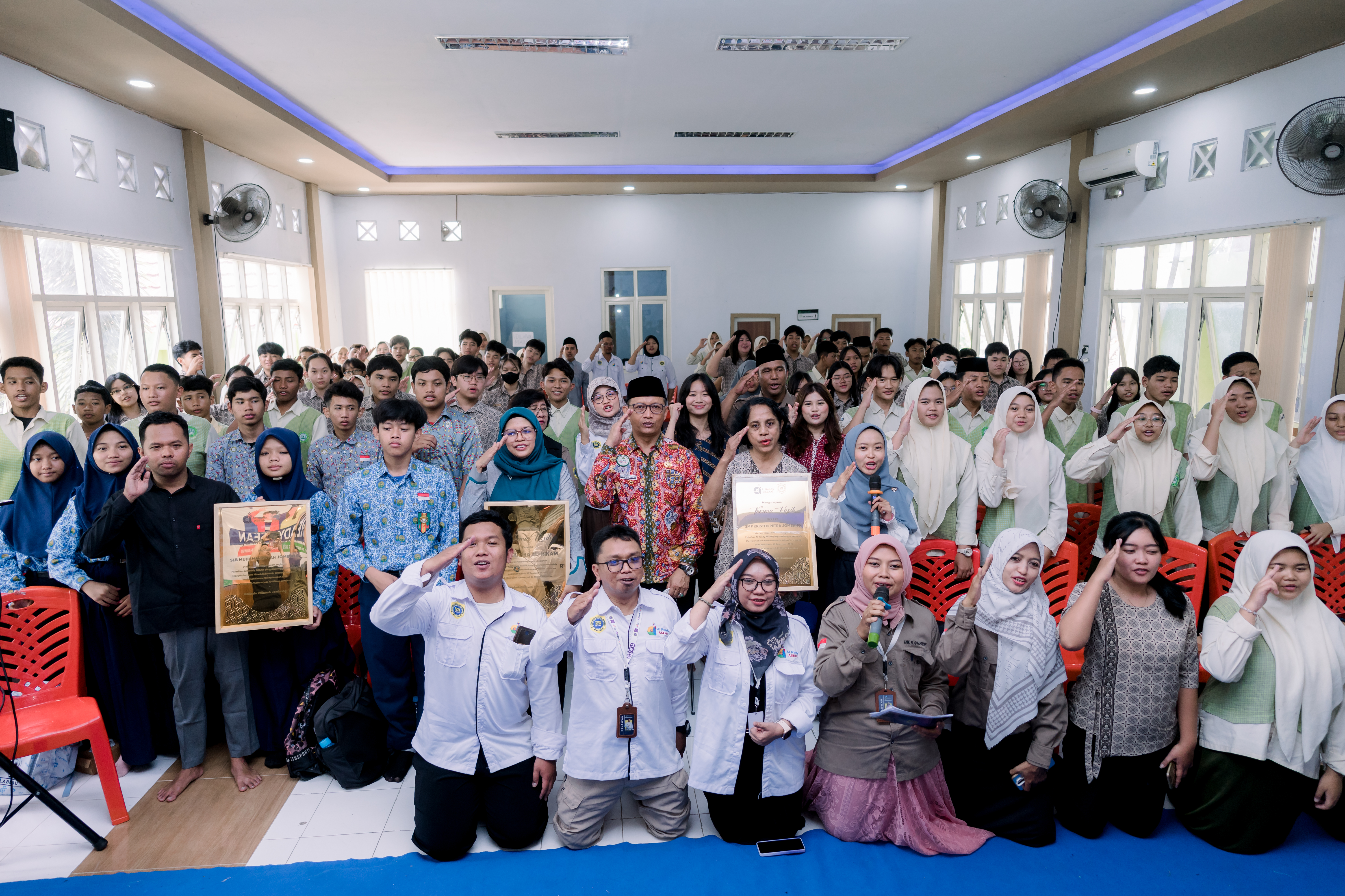 A large group photo of students and adults gathered in a brightly lit auditorium for the AI Ready ASEAN program. The group is posing, with many people giving a salute gesture. In the front row, adults hold up two framed plaques. The diverse crowd includes male and female participants.