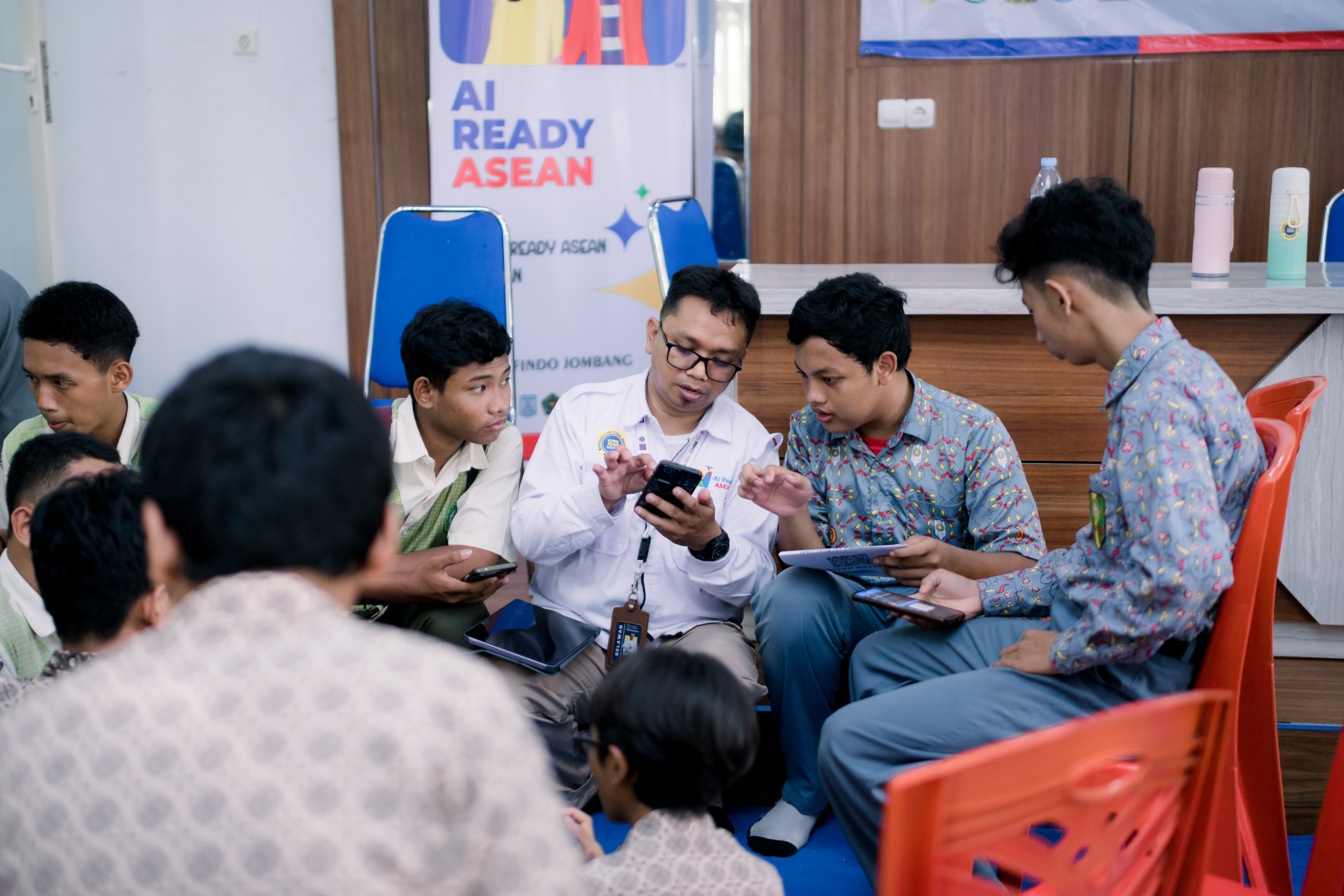 A man in a white shirt and glasses shows his smartphone to a small group of male students gathered around him. A backdrop behind them reads "AI READY ASEAN"
