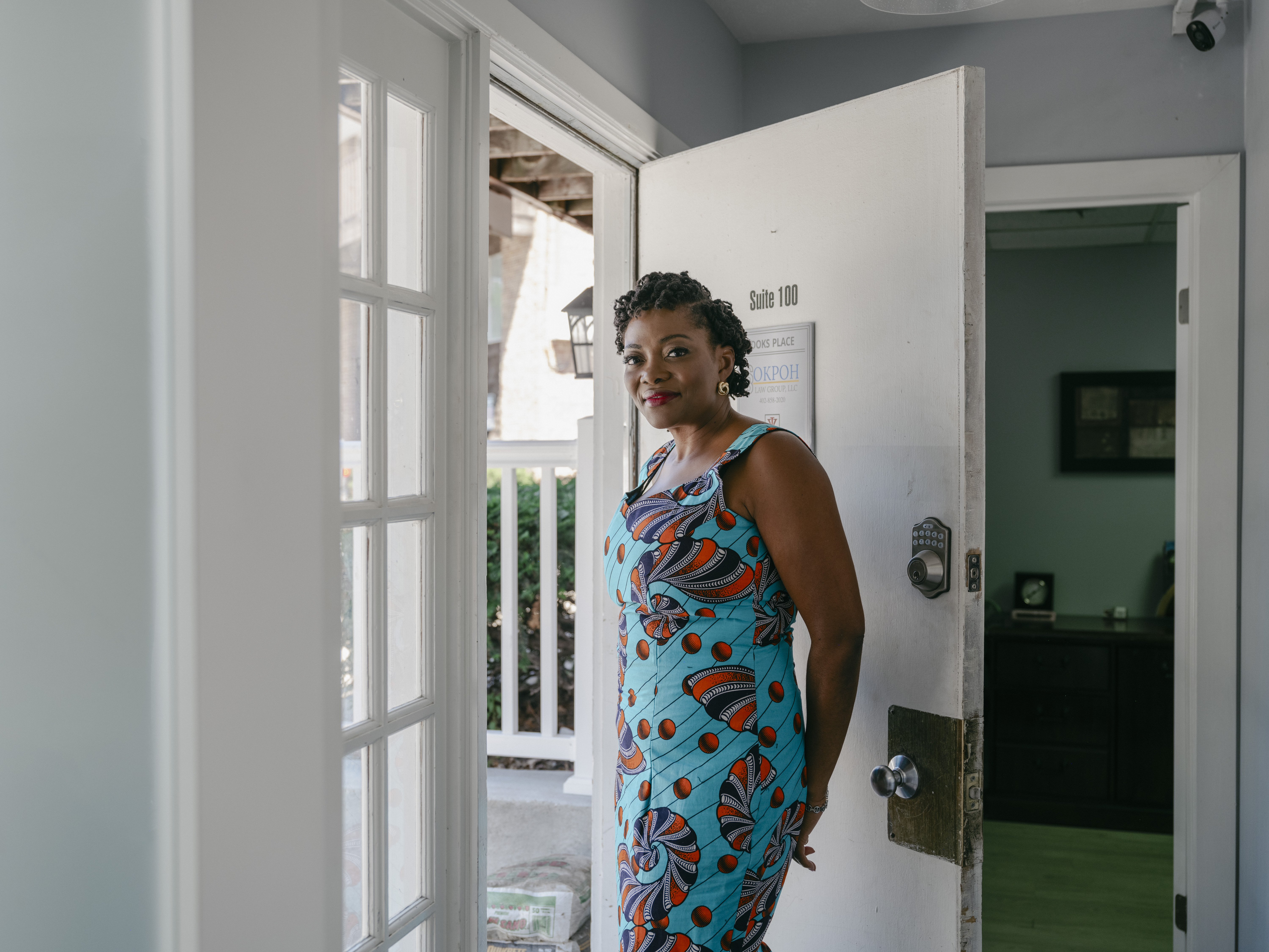 Karine Sokpoh, wearing a blue dress with a red and black pattern, stands in the doorway of the business she founded, Midlands African Chamber (MAC) in Omaha, Nebraska. Beyond the doorway is an office with a desk. To the left of Karine is a bank of small paned windows.