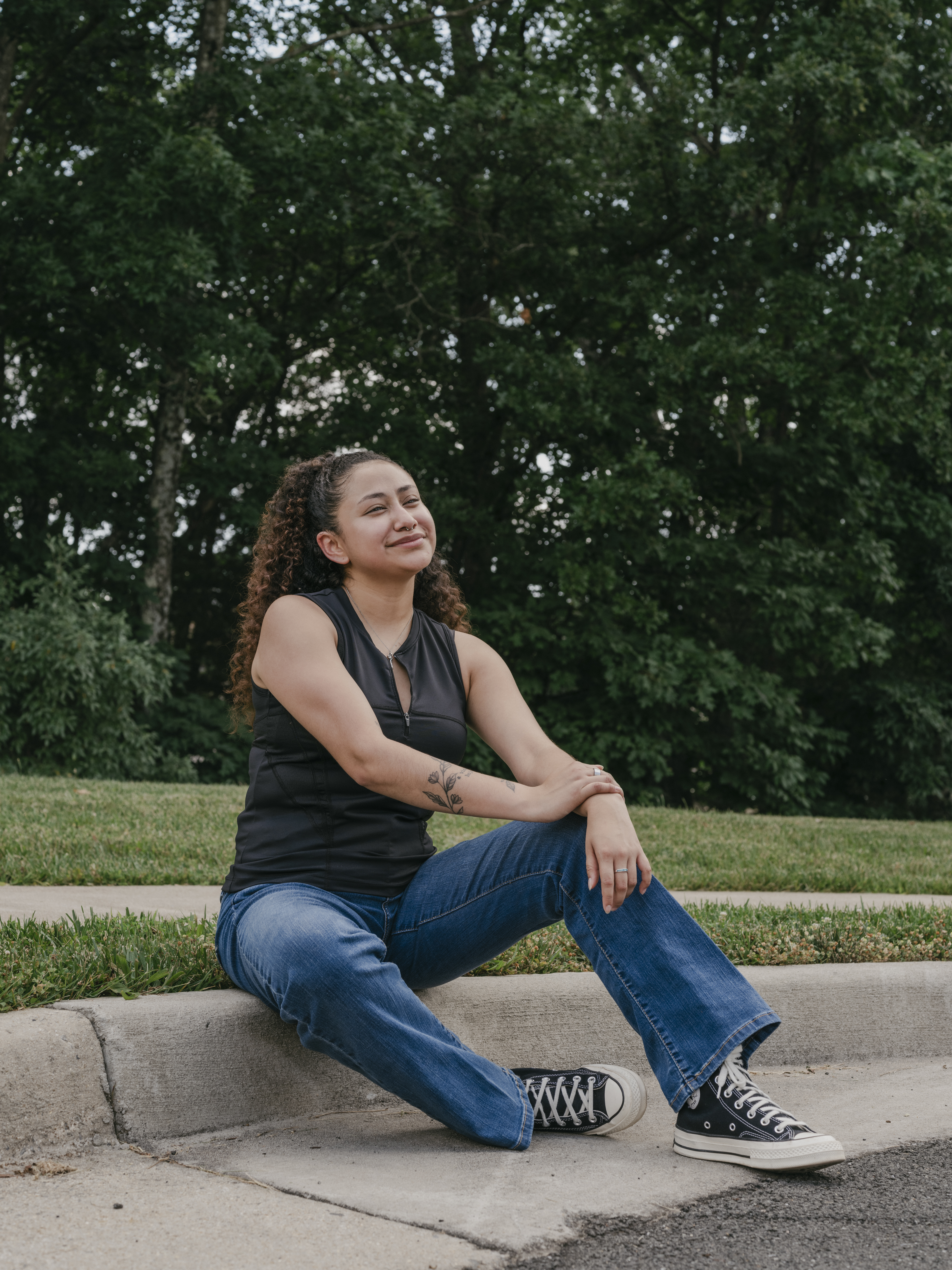 Aurora Rodriguez sits on a curb with one arm on her knee and the other arm laying on top of her opposite wrist. She is wearing a black top, blue jeans, and black and white sneakers. She smiles and her eyes are shut. Trees and shrubs are in the background.