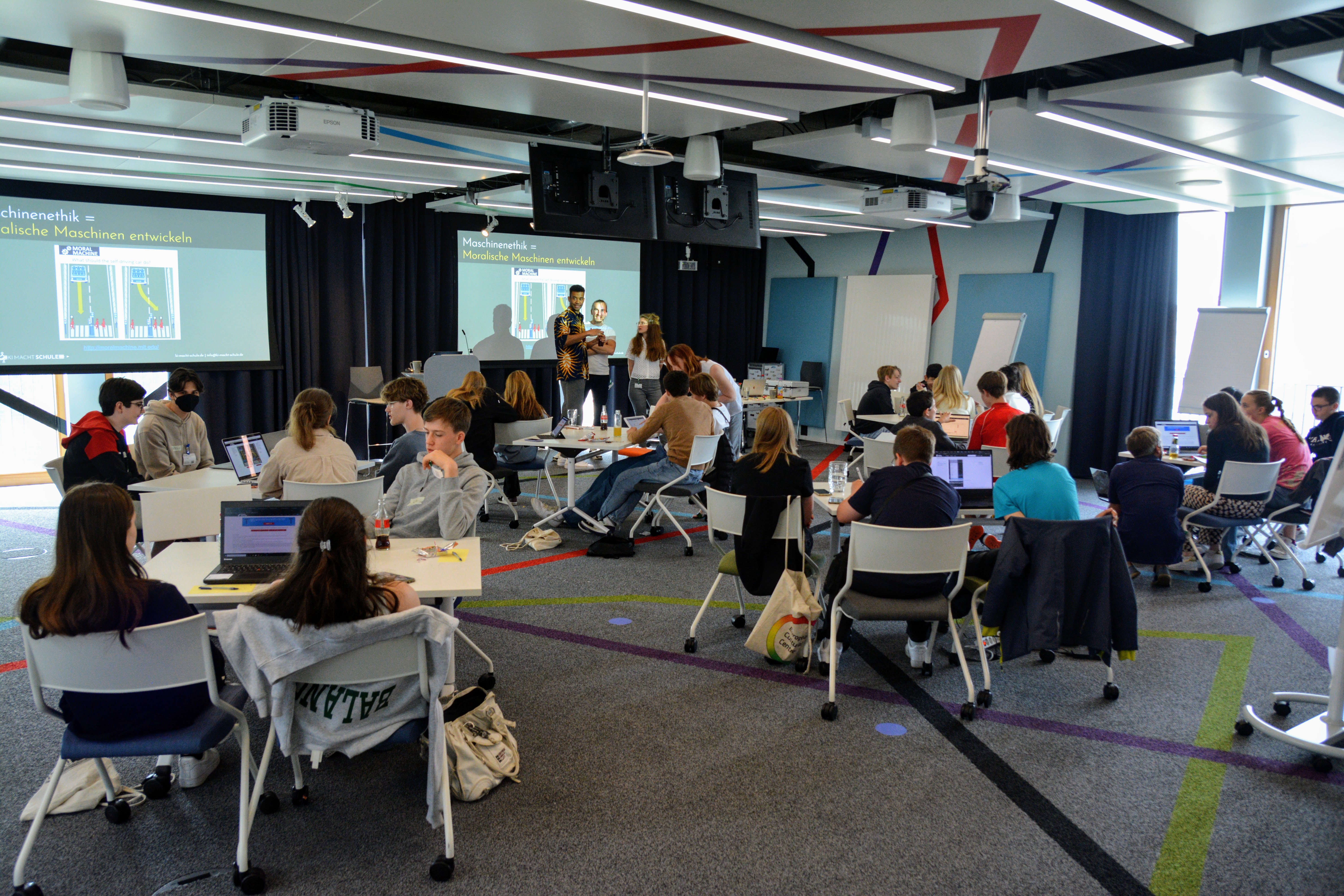 Koloriertes Foto auf dem eine Schülergruppe an Tischen sitzt, während ein Kursleiter zwischen zwei