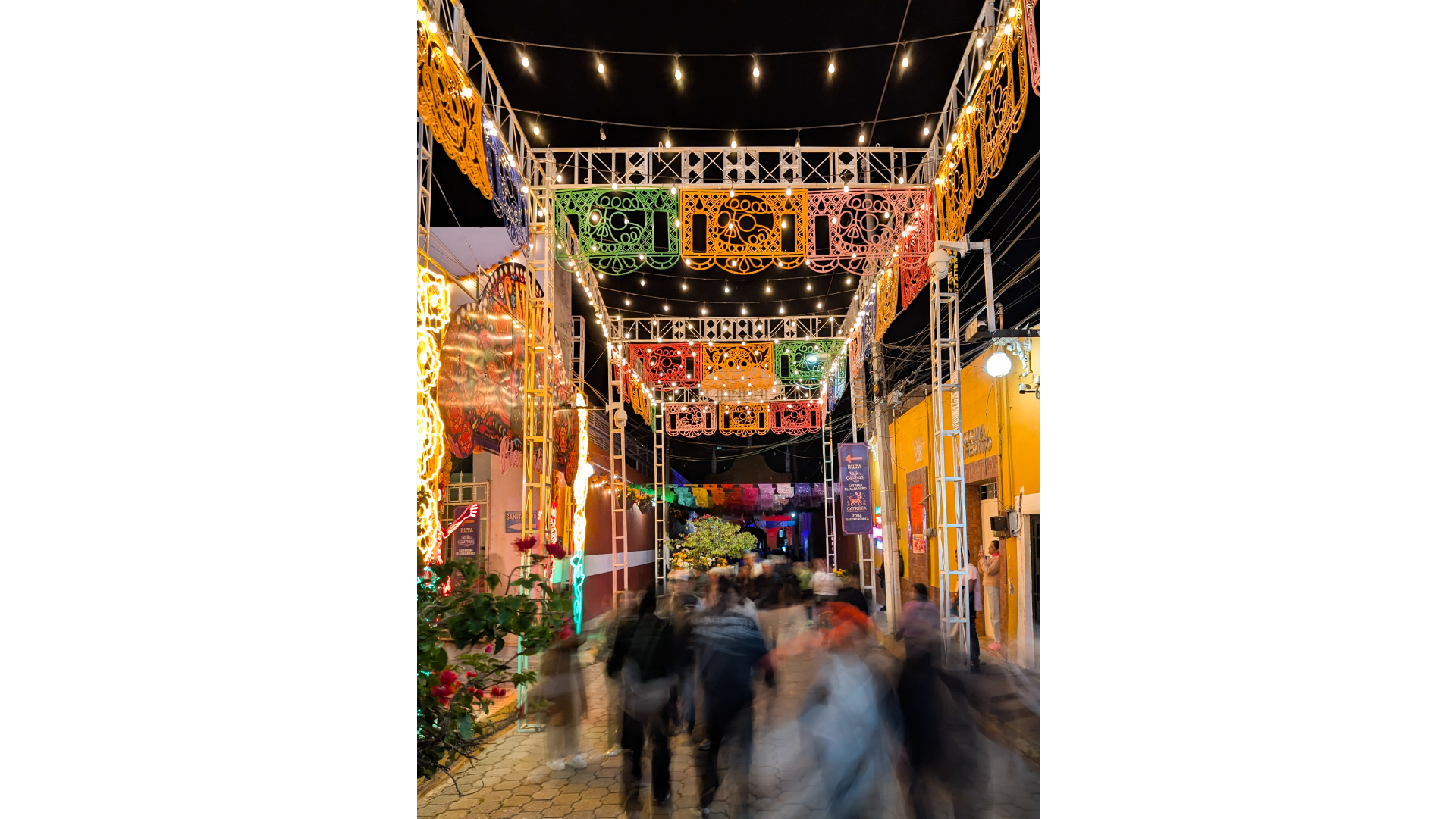 A long-exposure, nighttime photo of a festive street, where a blurred crowd walks under colorful papel picado banners and string lights.
