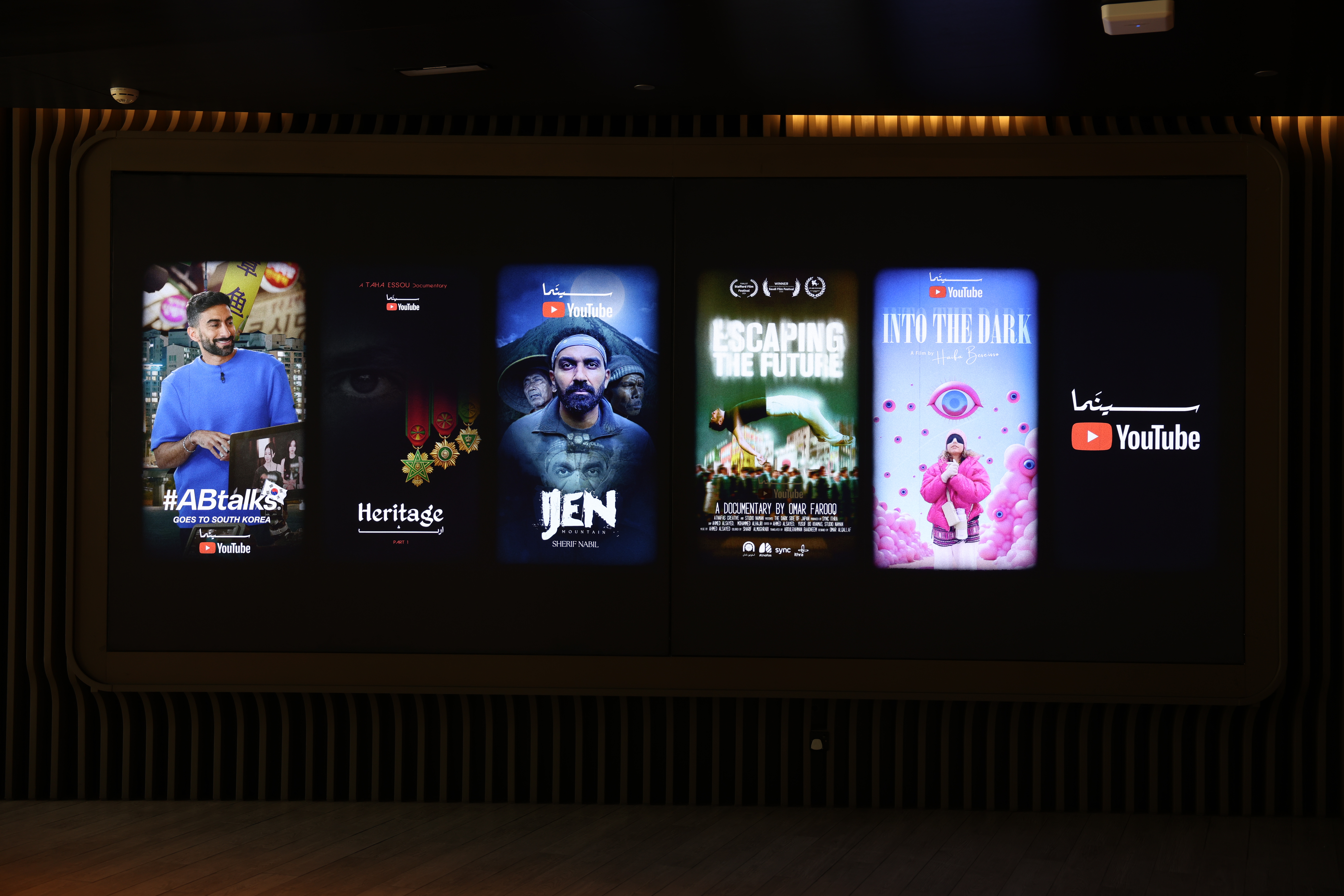 A large display screen shows four film posters, each presented within a smartphone-like shape, along with the "Cinema YouTube" logo. The first poster from the left is titled "#ABtalks" and features a man in a blue shirt. The second poster is titled "Heritage" and shows a close-up of a man's face. The third poster is titled "Jen" and features a man looking forward. The fourth poster is titled "Escaping The Future" and shows a green building. The last poster, on the far right, is titled "Into The Dark" and shows a person wearing a large pink coat. All posters are accompanied by the YouTube logo.