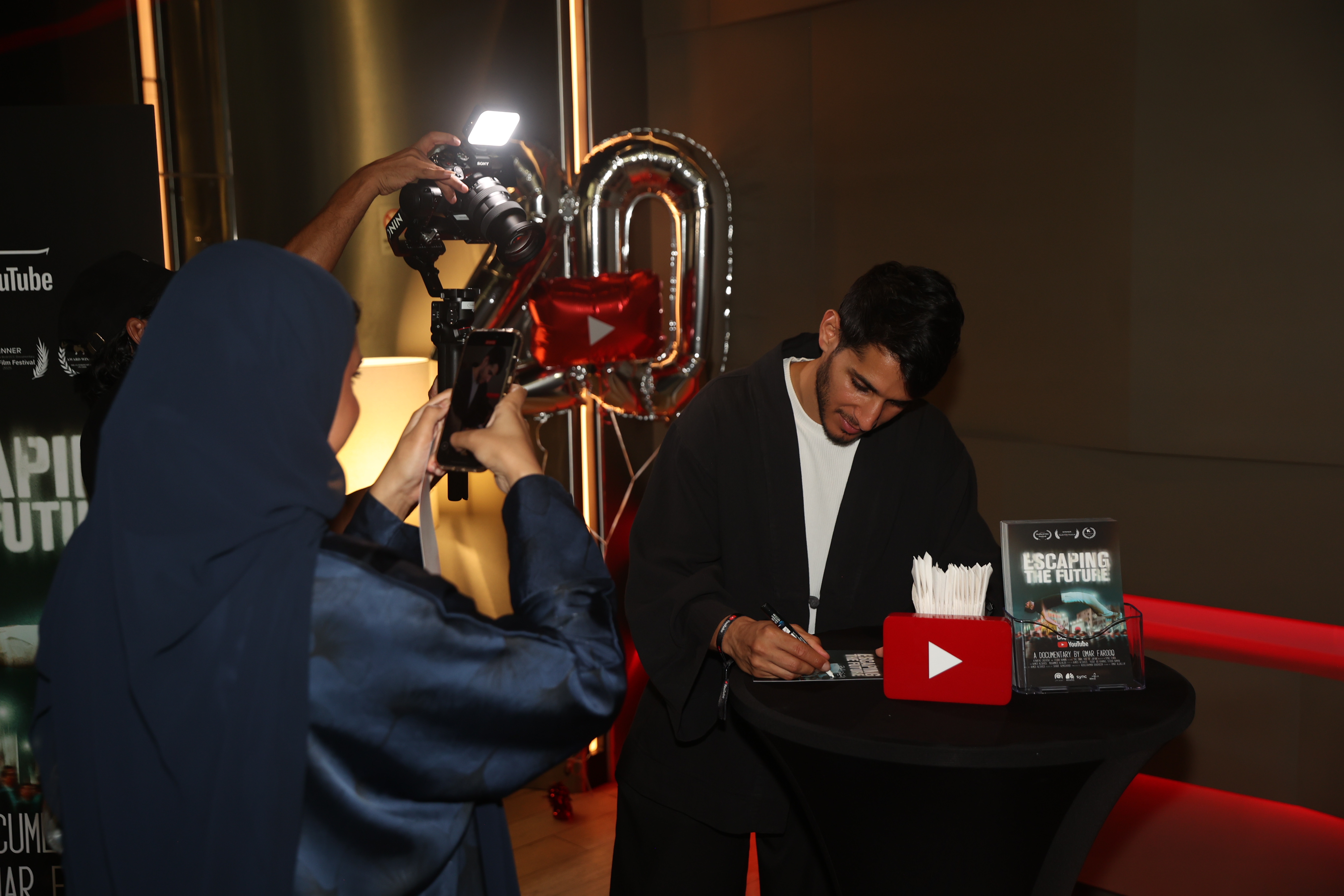 A young man wearing a black jacket over a white shirt signs a poster for the film "Escaping the Future" placed on a small black table. Next to him is a small display stand holding a red YouTube logo model. Behind the man, large silver balloons forming the number "20" and a small red YouTube logo balloon are visible. On the left side of the image, a woman wearing a blue hijab films the man with her phone, while another photographer with a professional camera stands behind her. Parts of a "YouTube" banner are visible on the wall in the background.