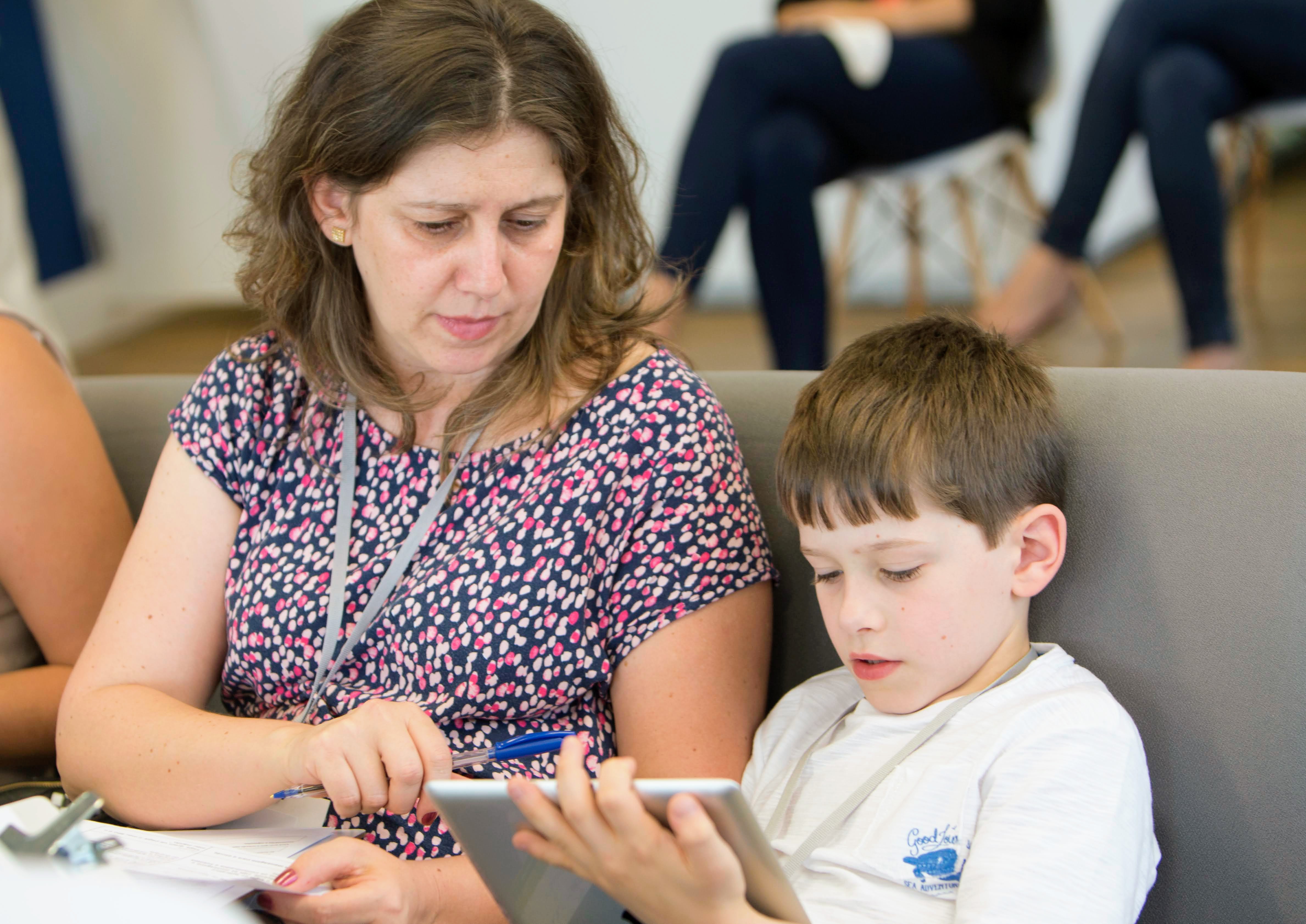 Woman and boy view a tablet he holds; she holds a pen and papers.