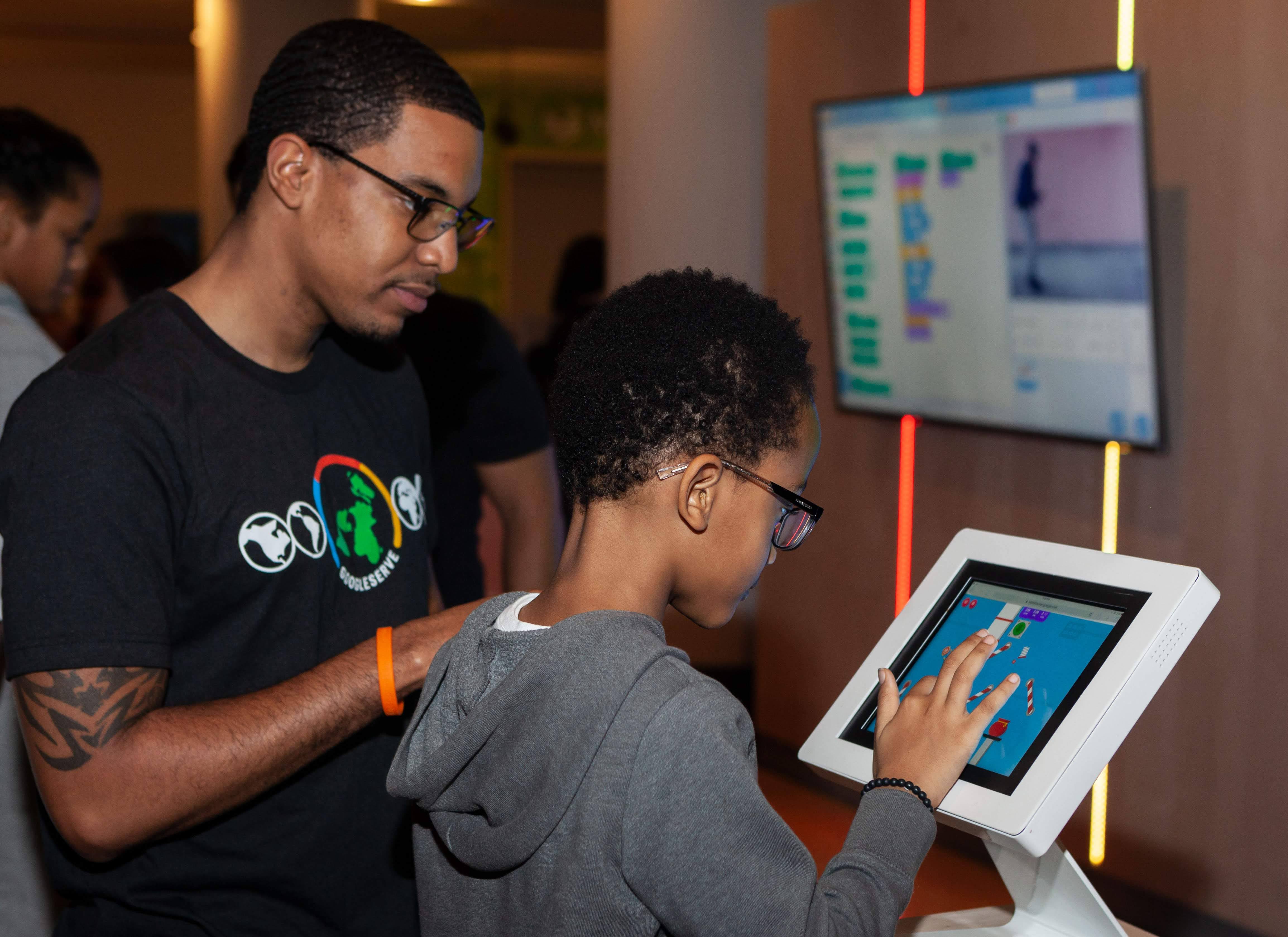 Child taps tablet game, man observes; programming code in background.