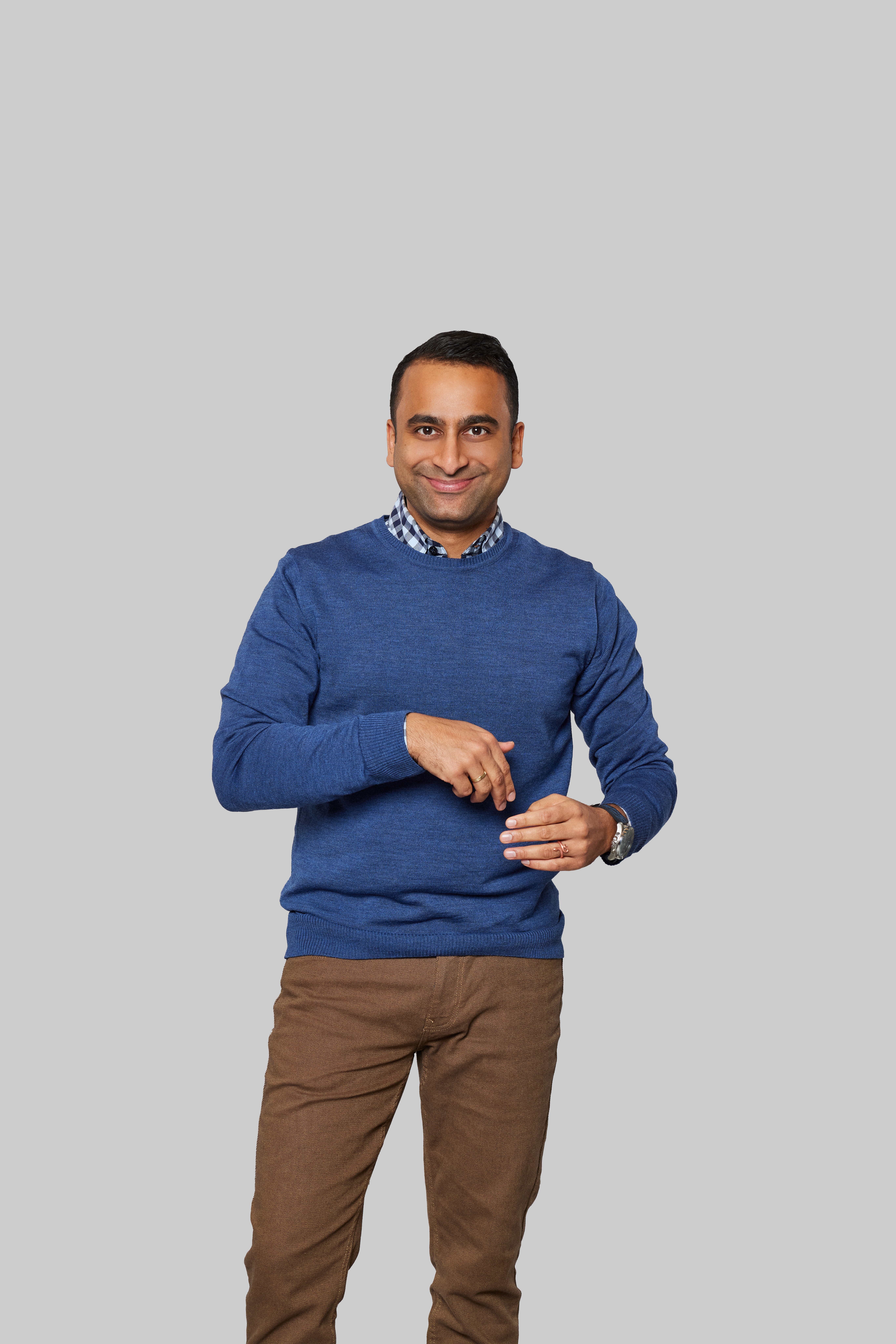 Na zdjęciu stoi mężczyzna na szarym tle. Jest uśmiechnięty, ubrany w niebieski sweter i brązowe spodnie, na lewej ręce ma zegarek.