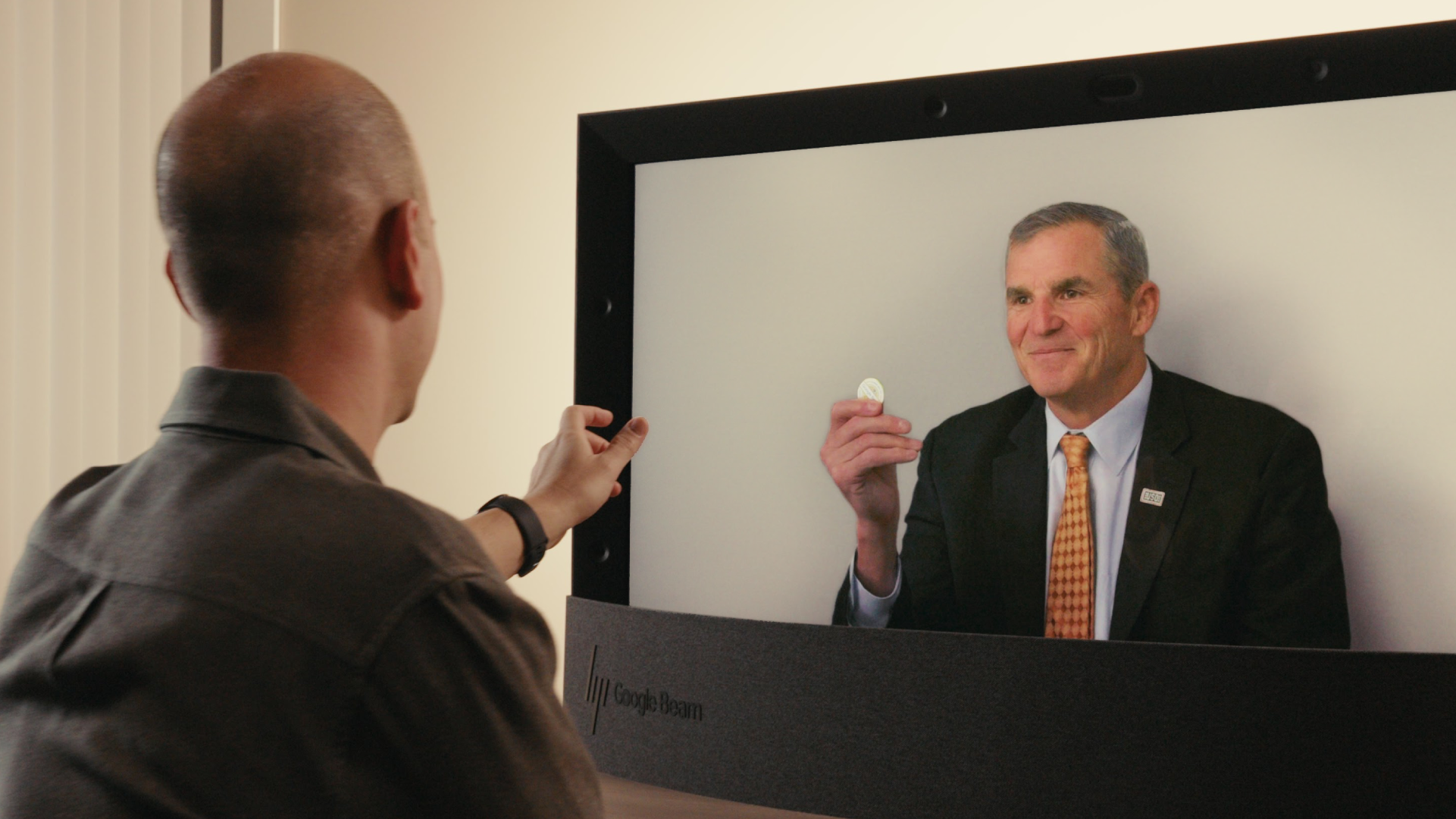 This is a static image for the Google Beam 3D video communication system, highlighting its ability to convey subtle interactions and handle objects with a realistic sense of depth. The image perfectly illustrates the Beam technology's ability to render objects and gestures with a realistic sense of dimension. The USO CEO is holding the coin up to the camera, and the depth of the image makes it appear as though the coin is being presented directly across the desk to the local participant.