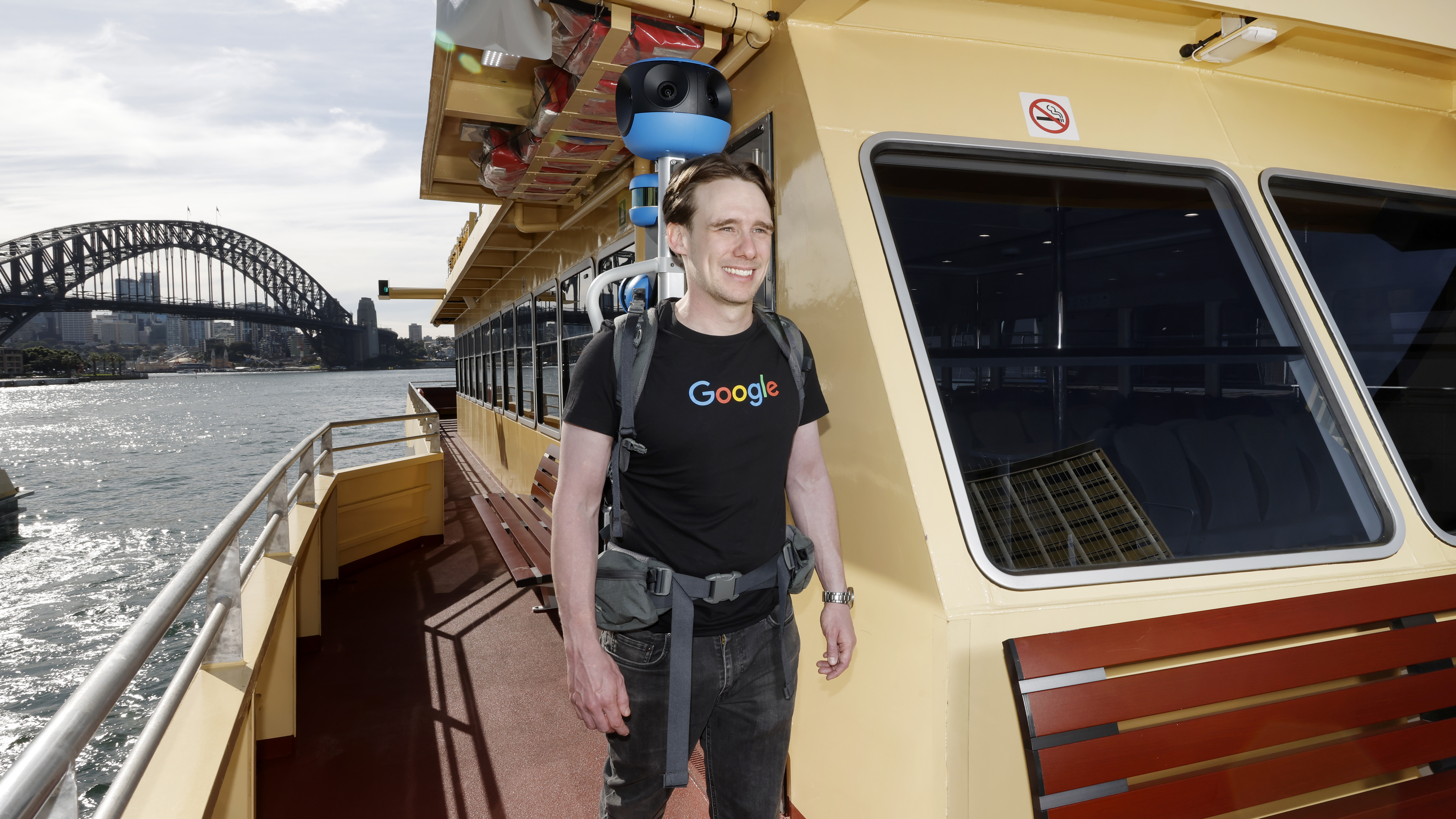 A photo showing Daniel Nadasi walking down the side of the outside deck in a green and yellow ferry, wearing the blue Street View trekker backpack.