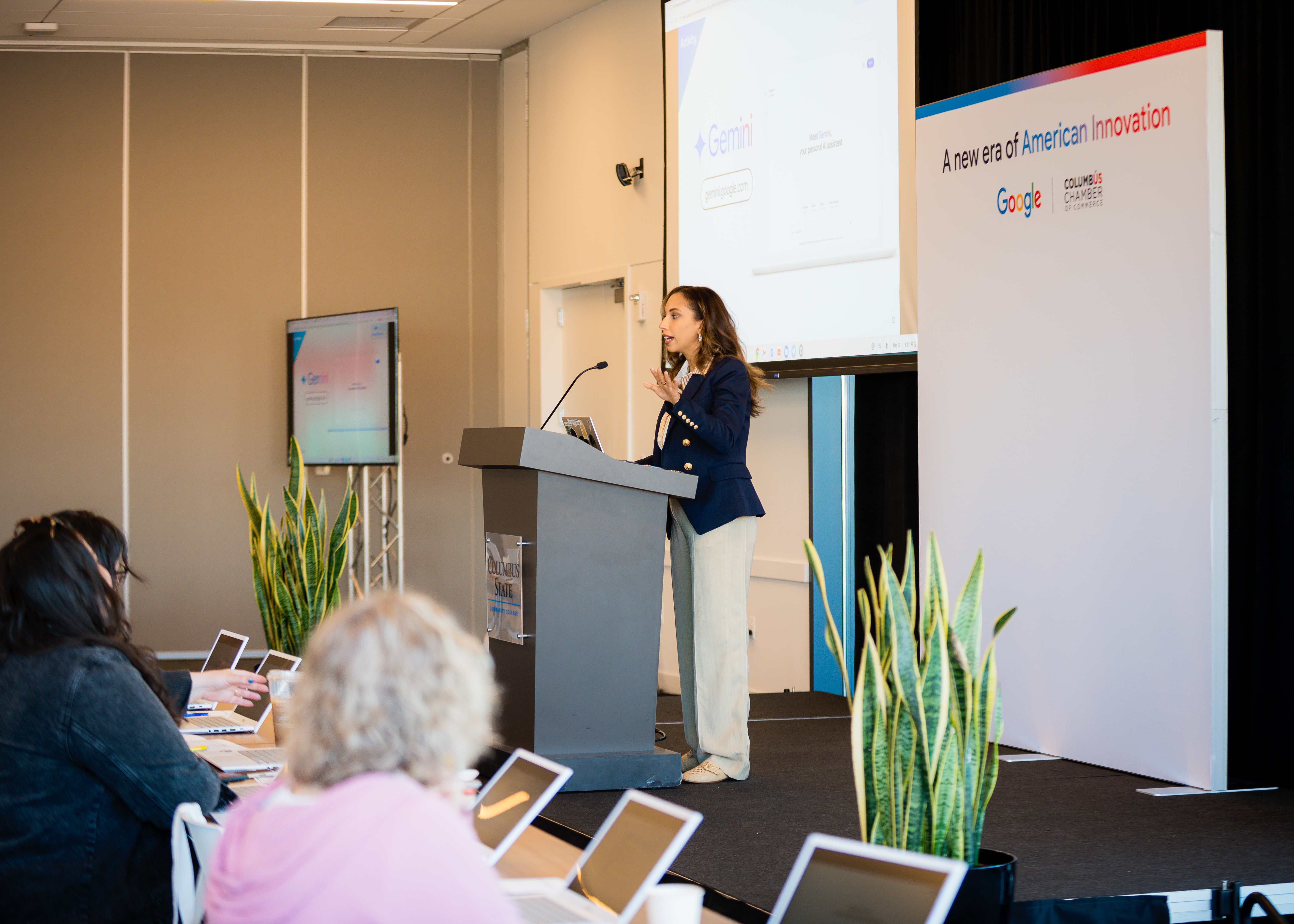 Woman speaking at podium, 'Gemini' on screen, 'American Innovation' banner.
