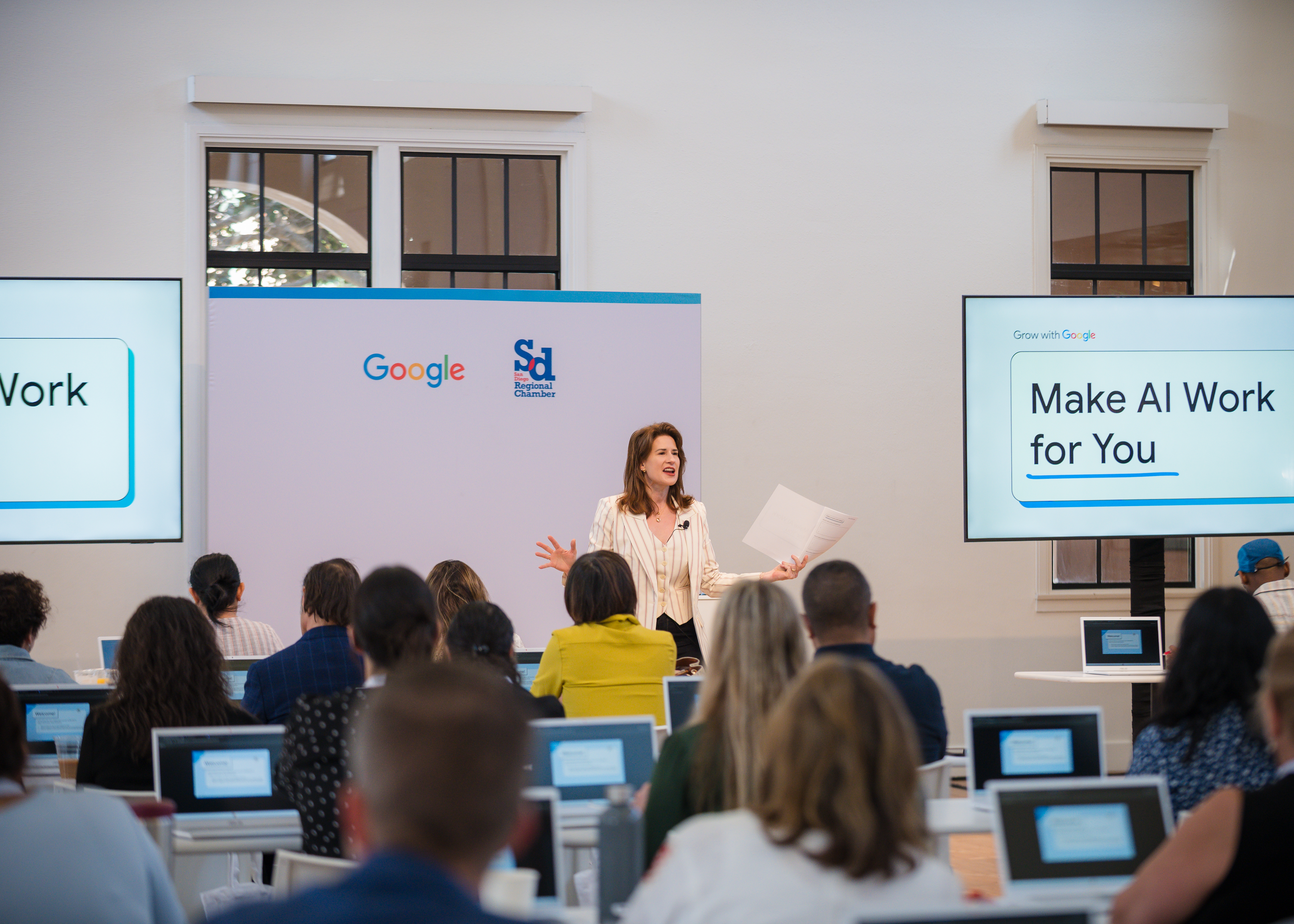 Woman speaks before screens displaying Google and San Diego Regional Chamber logos, and "Make AI Work for You."