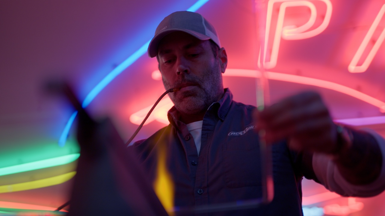 Photo of a man working on something with his hands. The photo is slightly blurry; he is backlit by neon lights behind him.