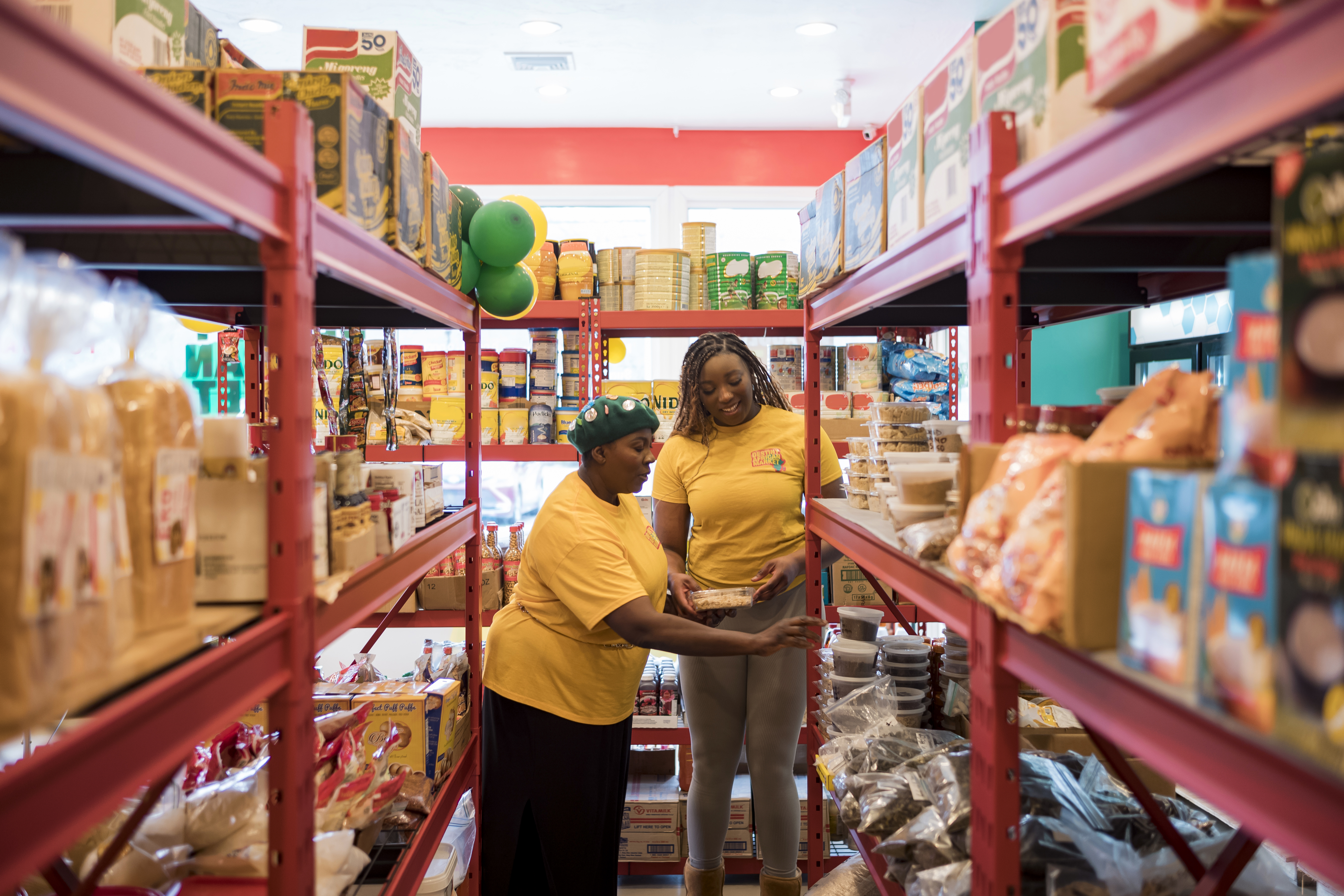 Owner Sola and a colleague check stock at their market. They’re flanked by red shelves full of products extending towards the camera.