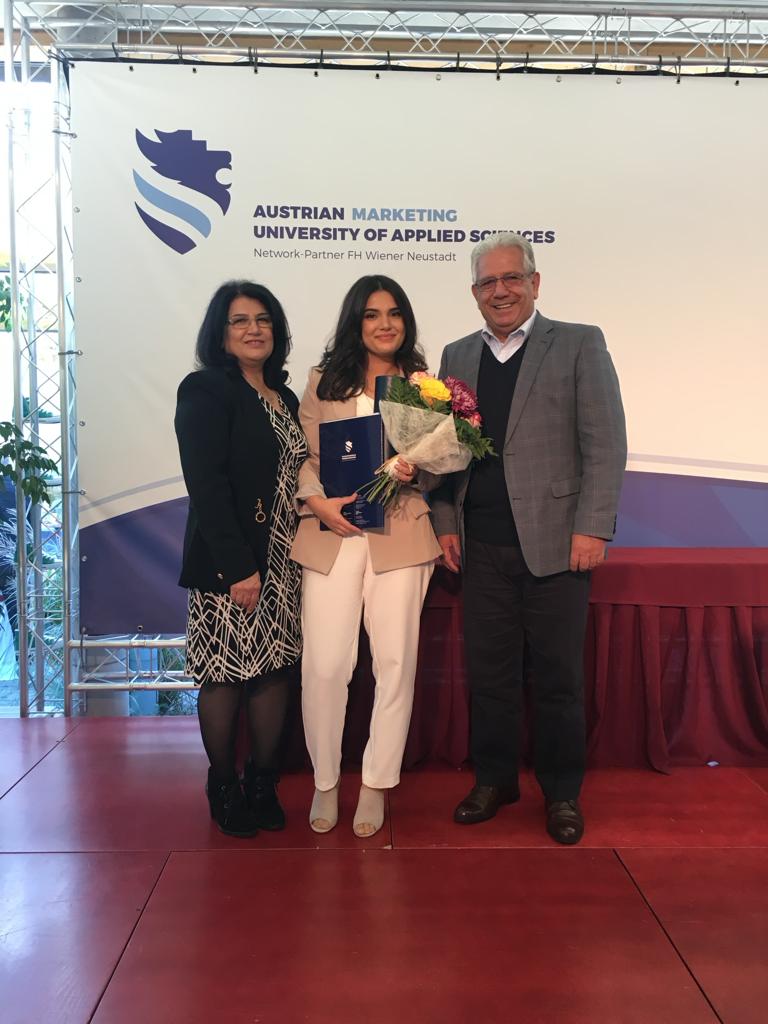 Googlerin Maryam Hakimi steht vor einer Logowand der „Austrian Marketing University of Applied Siences“ und häält stolz ihren Masterabschluss und einen Strauß Blumen in der Hand. Links von ihr steht ihr Vater, rechts ihre Mutter.