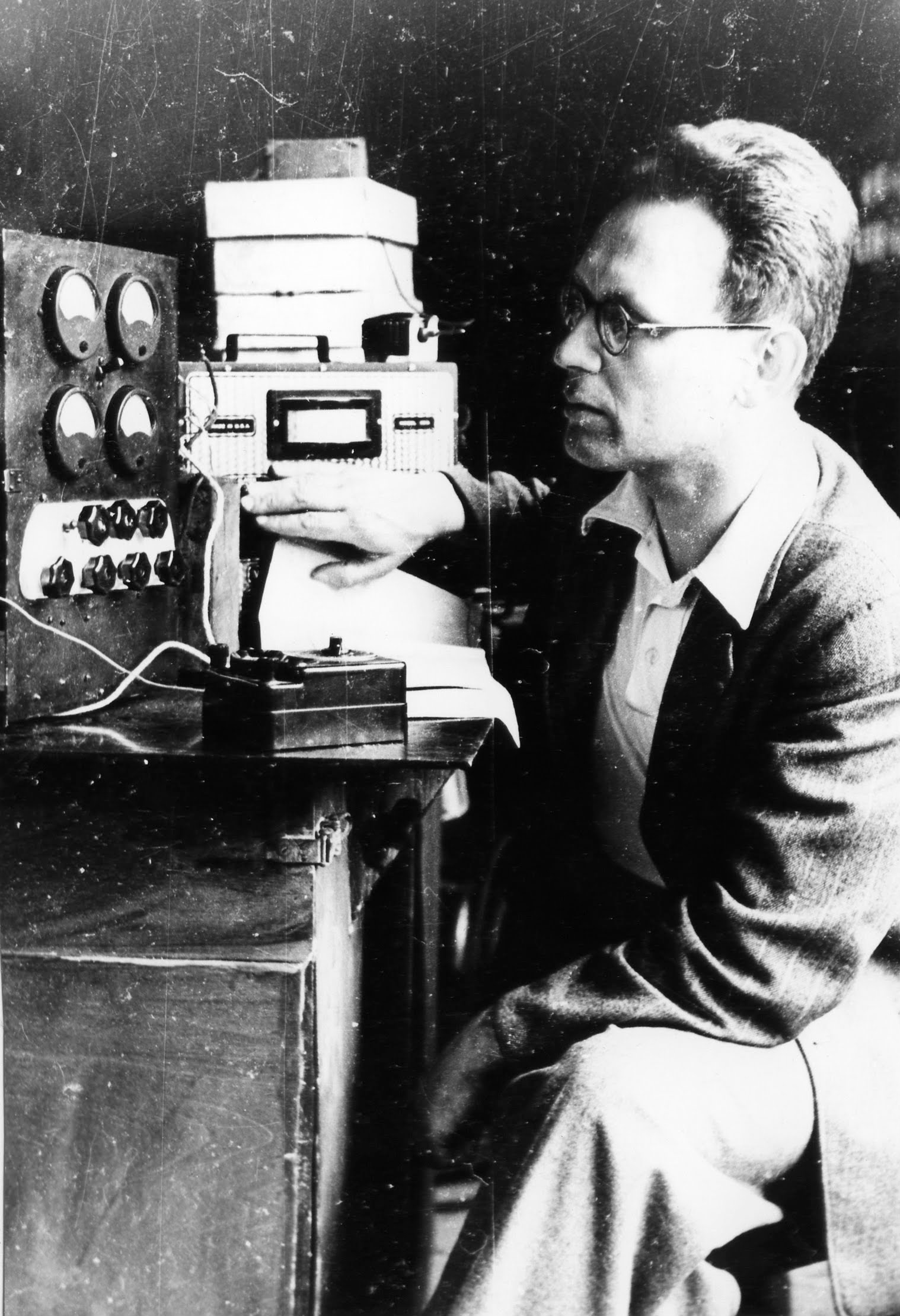 A black and white photo of a spectacled man sitting beside a machine.