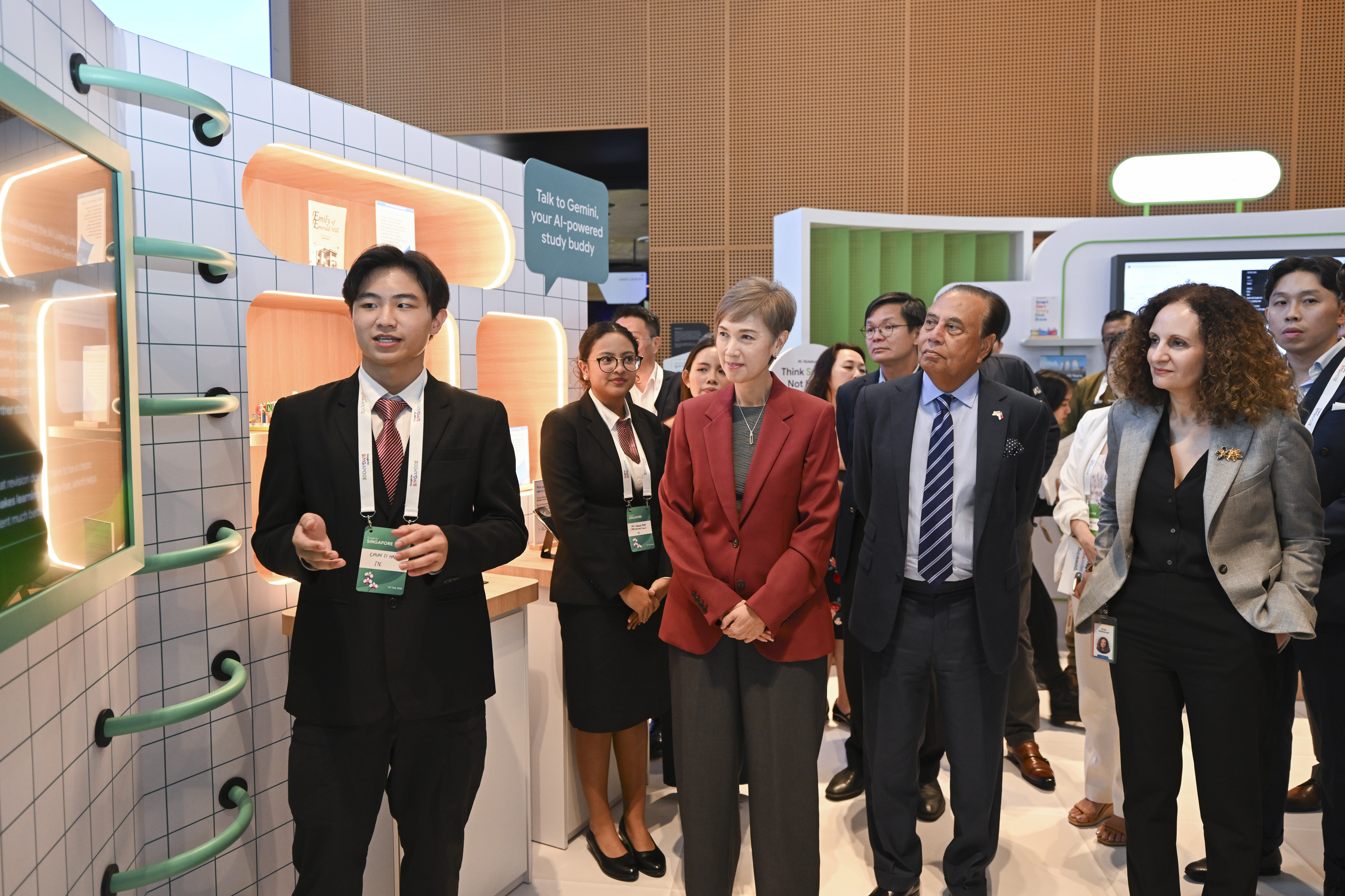 A young presenter in a suit gestures toward a digital display while speaking to a group of officials, including a woman in a red blazer. Behind them is a booth with the text "Talk to Gemini, your AI-powered study buddy" and a light-up graphic.