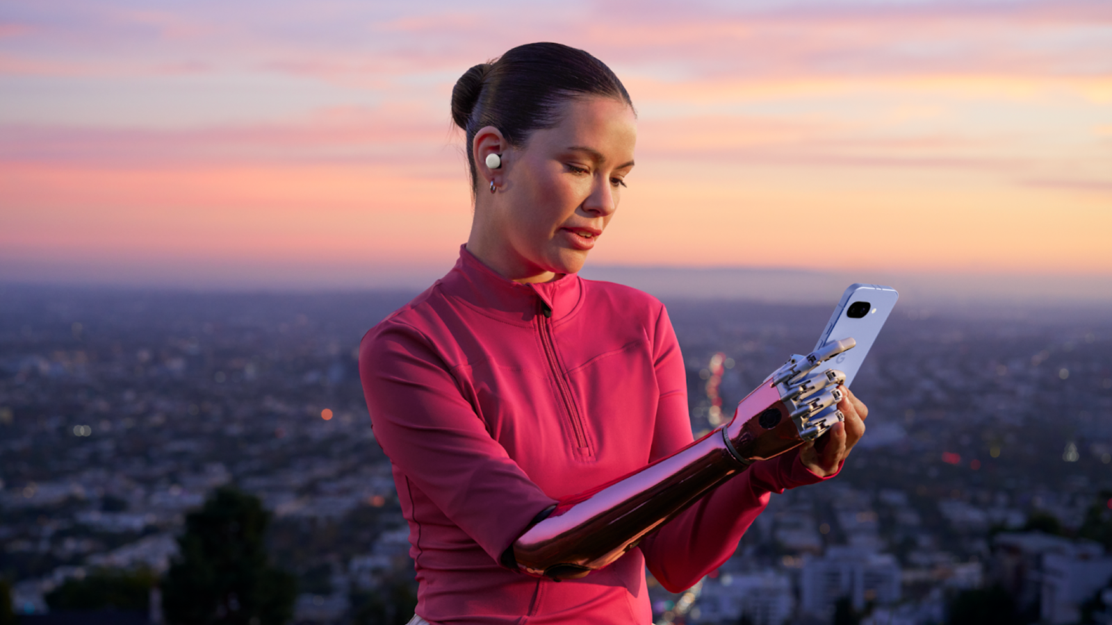 A person is looking down at a light-colored smartphone they are holding. They have a prosthetic arm. The person has a wireless earbud in their left ear. The background is a blurred cityscape at dusk or dawn with a colorful sky.
