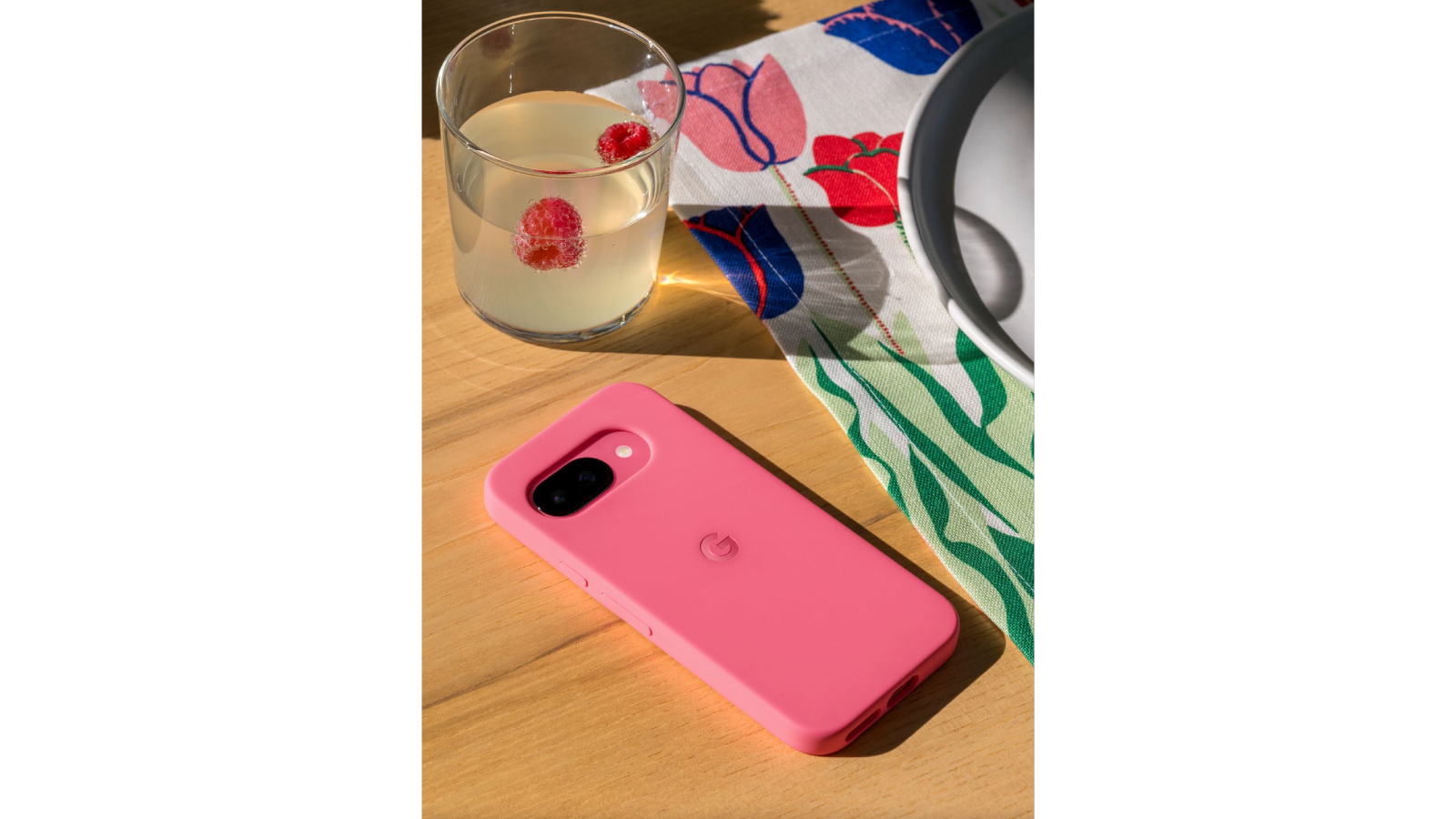 A bright pink smartphone in a matching case rests on a light brown wooden surface. The phone displays a black camera module and a subtle G logo in the center. To the left, a clear glass filled with a light-colored drink and two raspberries sits partially in the frame.