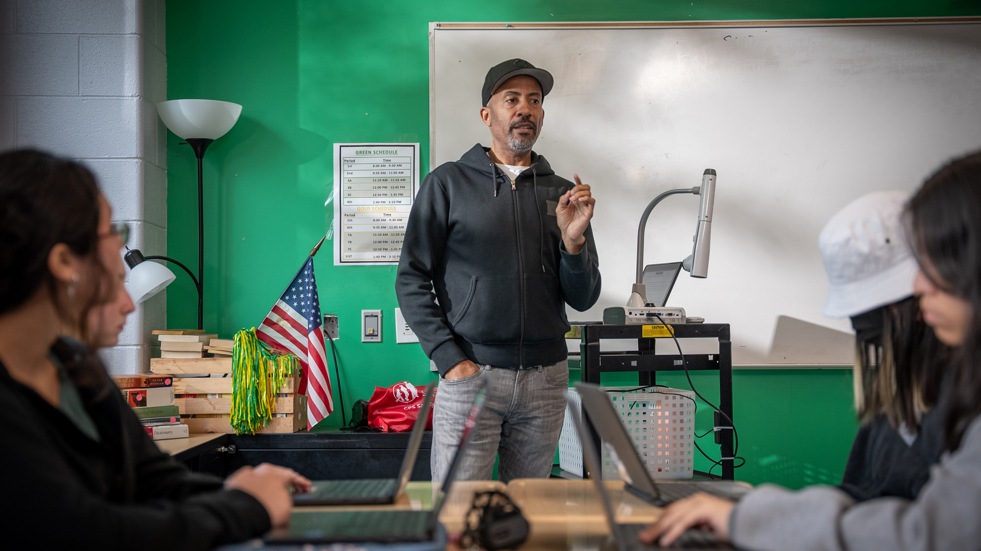 A teacher wearing a black hoodie, gray pants, and a black baseball cap stands at the front of a classroom and addresses students. Four students sit in the foreground with laptop computers.