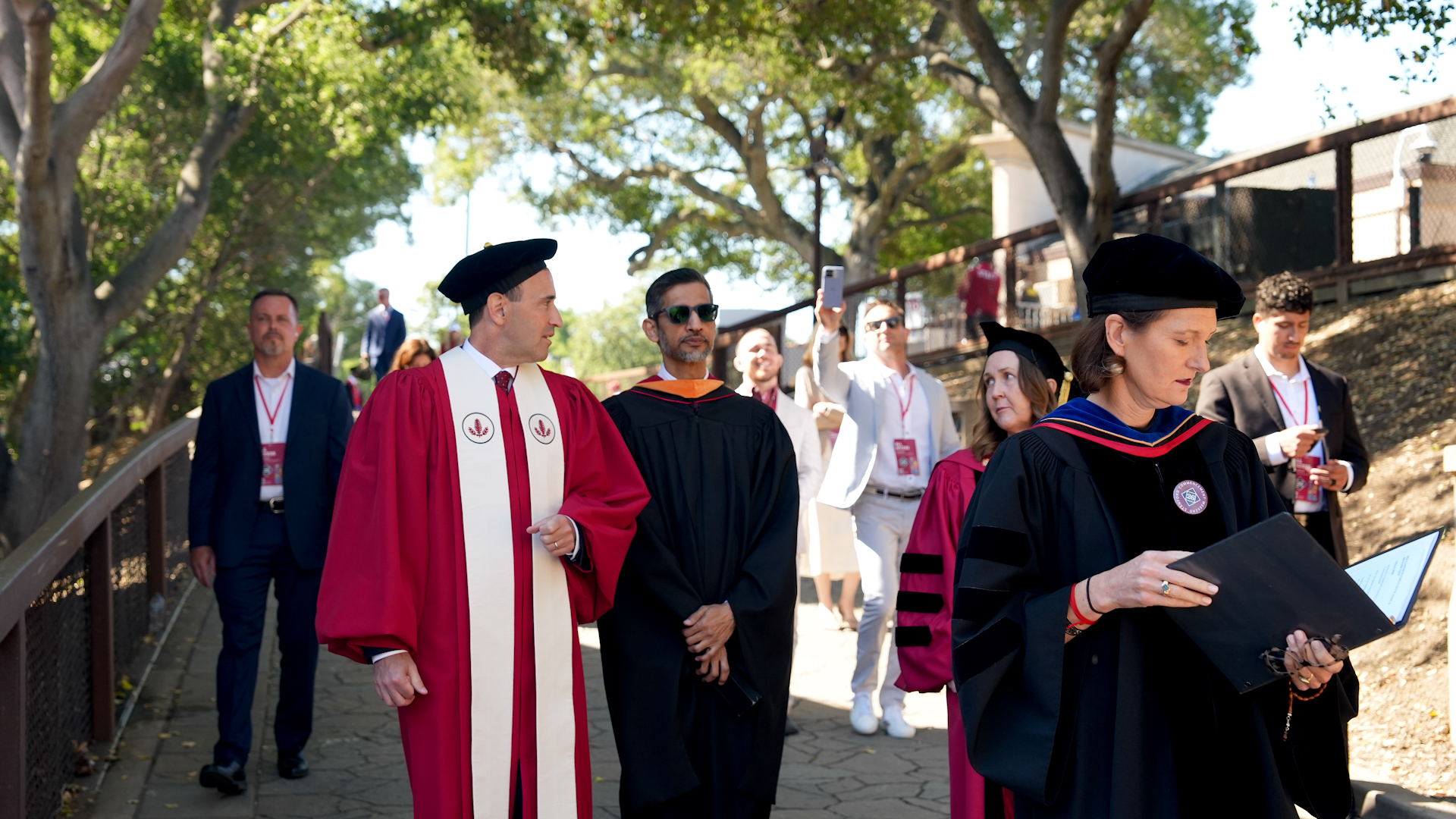 Academic procession in regalia walking along a sun-dappled, tree-lined path.