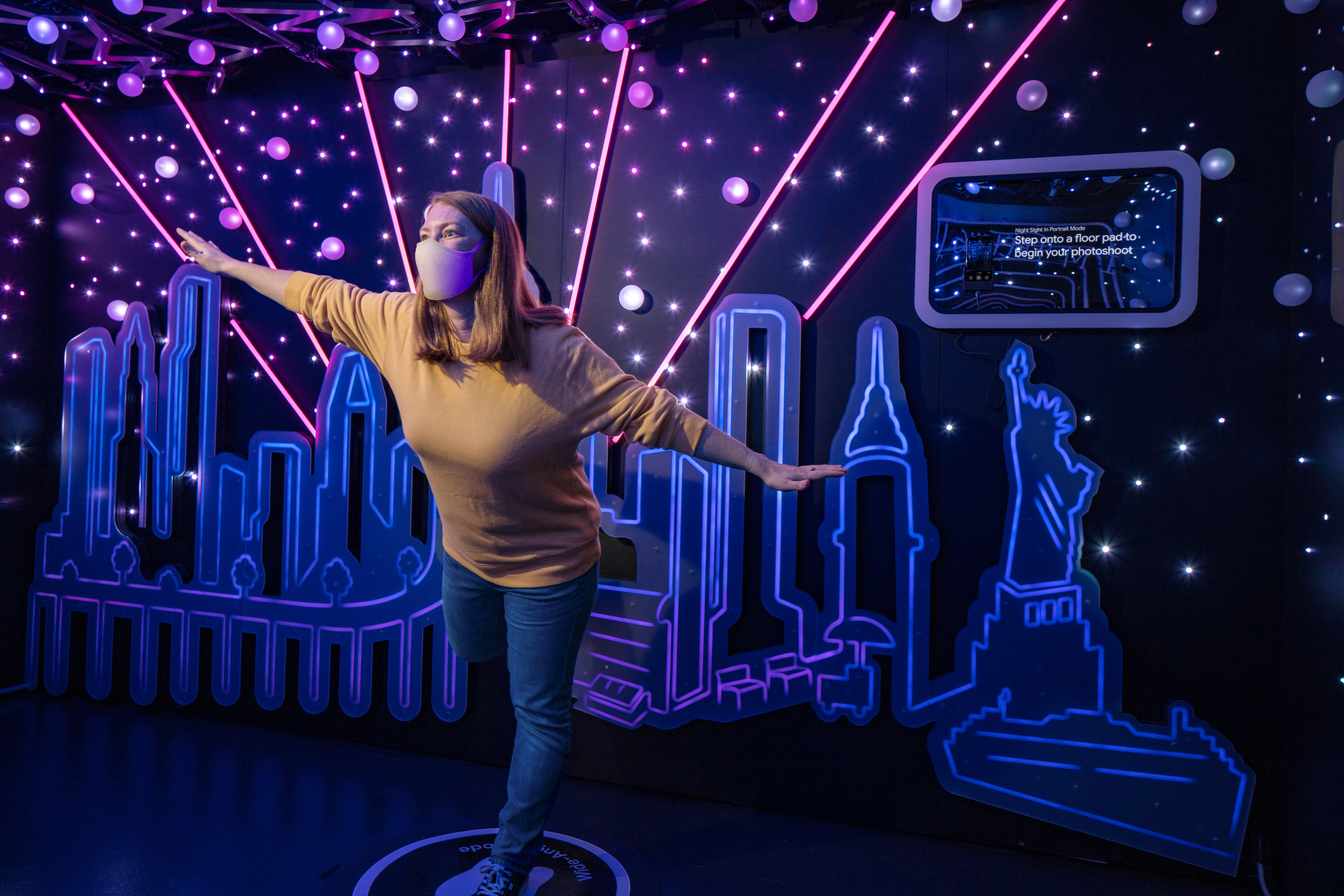 A person poses in front of a neon-lit New York cityscape in a dark room.