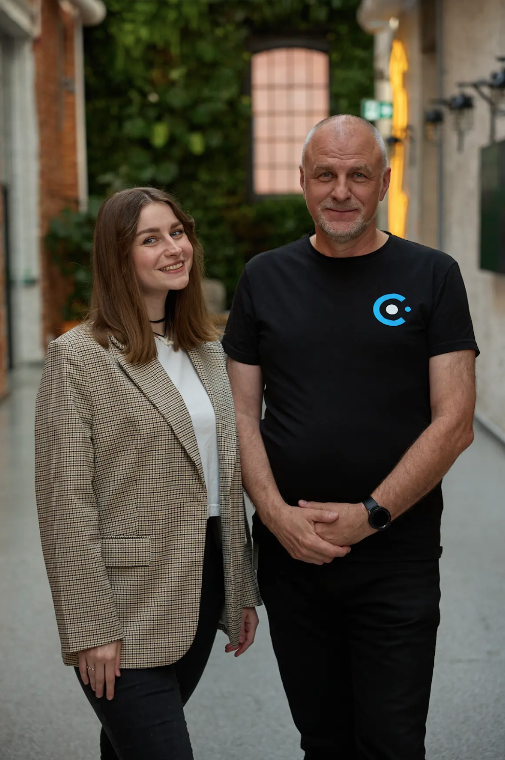 A woman in a tan blazer and a man in a black t-shirt stand side-by-side in a hallway.