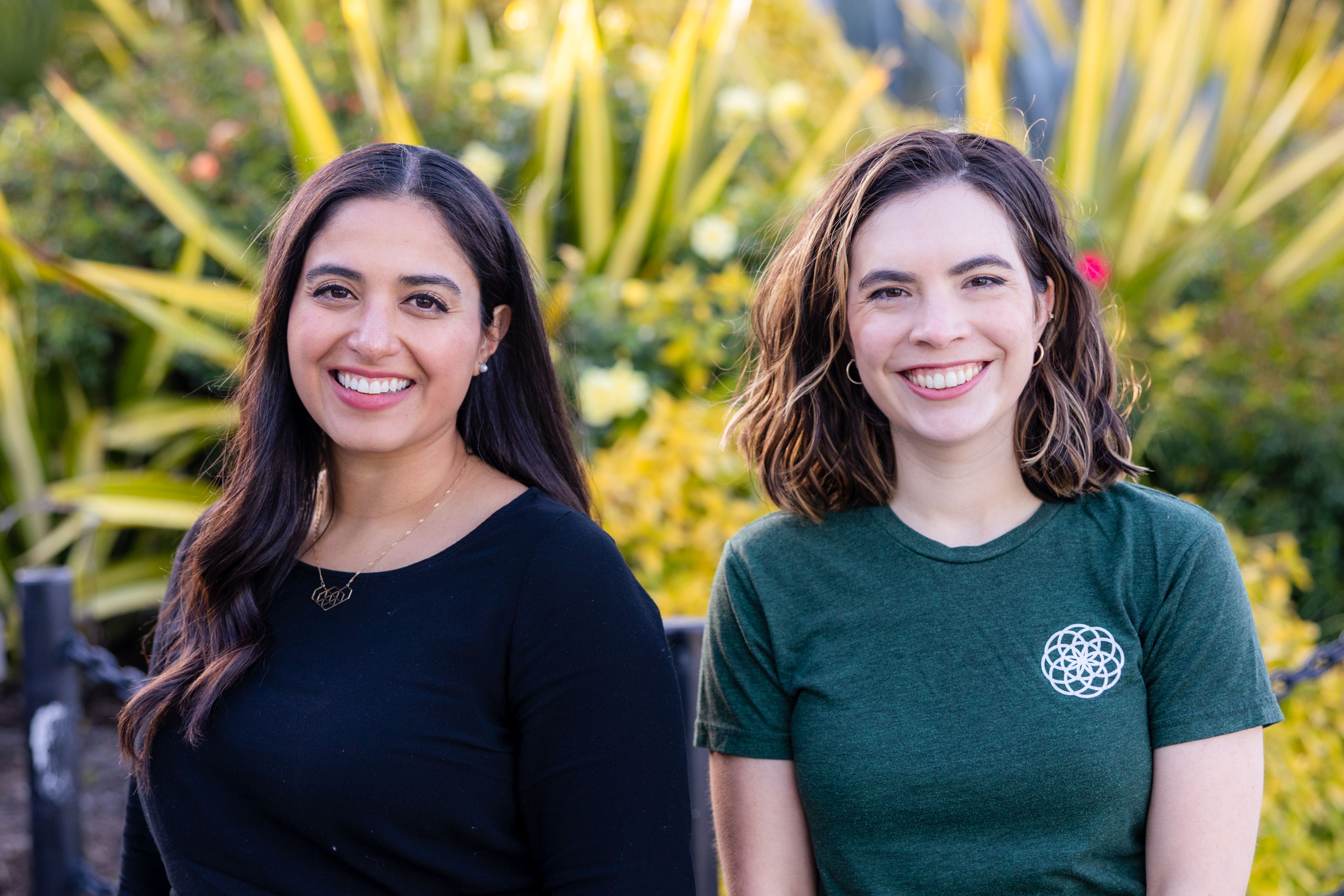 Two women, one wearing a black shirt and one wearing a green shirt, sit side by side facing the camera in front of large green plants.