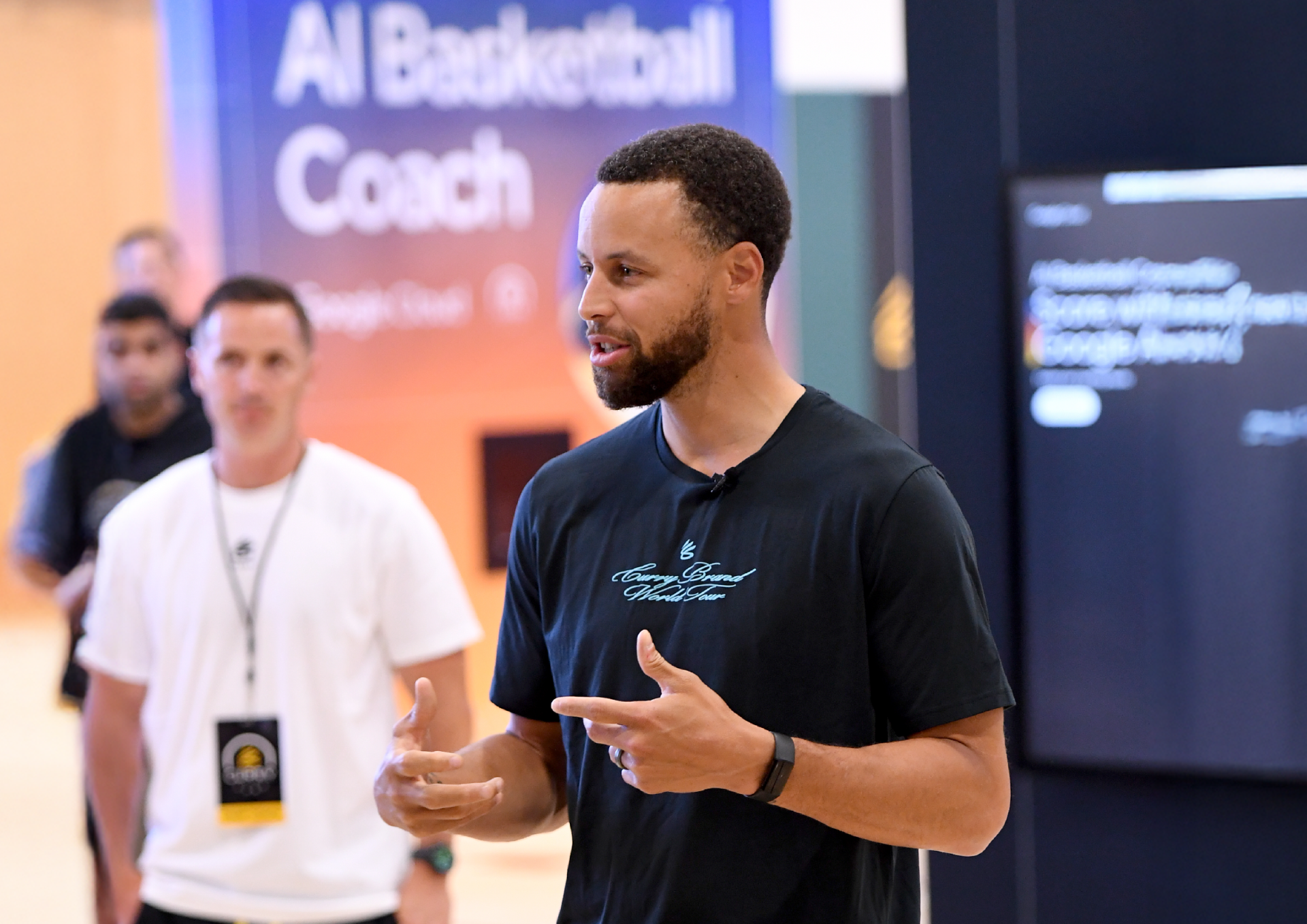 Stephen Curry standing wearing black tshirt; man with white tshirt and 'AI Basketball Coach' display in the background
