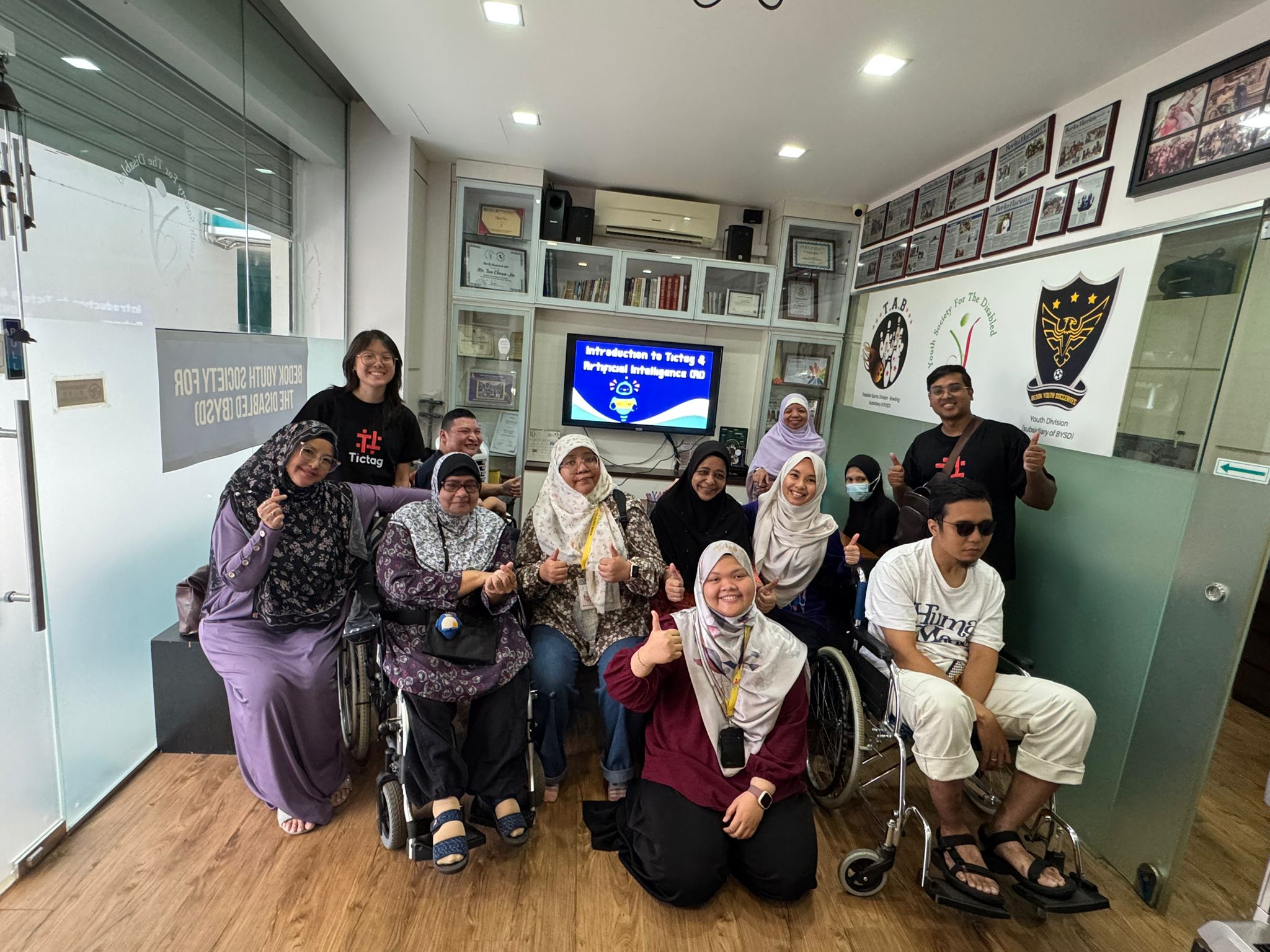 A group of diverse individuals, including several wheelchair users and women wearing hijabs, pose together for a photo in an office setting after an "Introduction to Tictag & Artificial Intelligence" presentation.