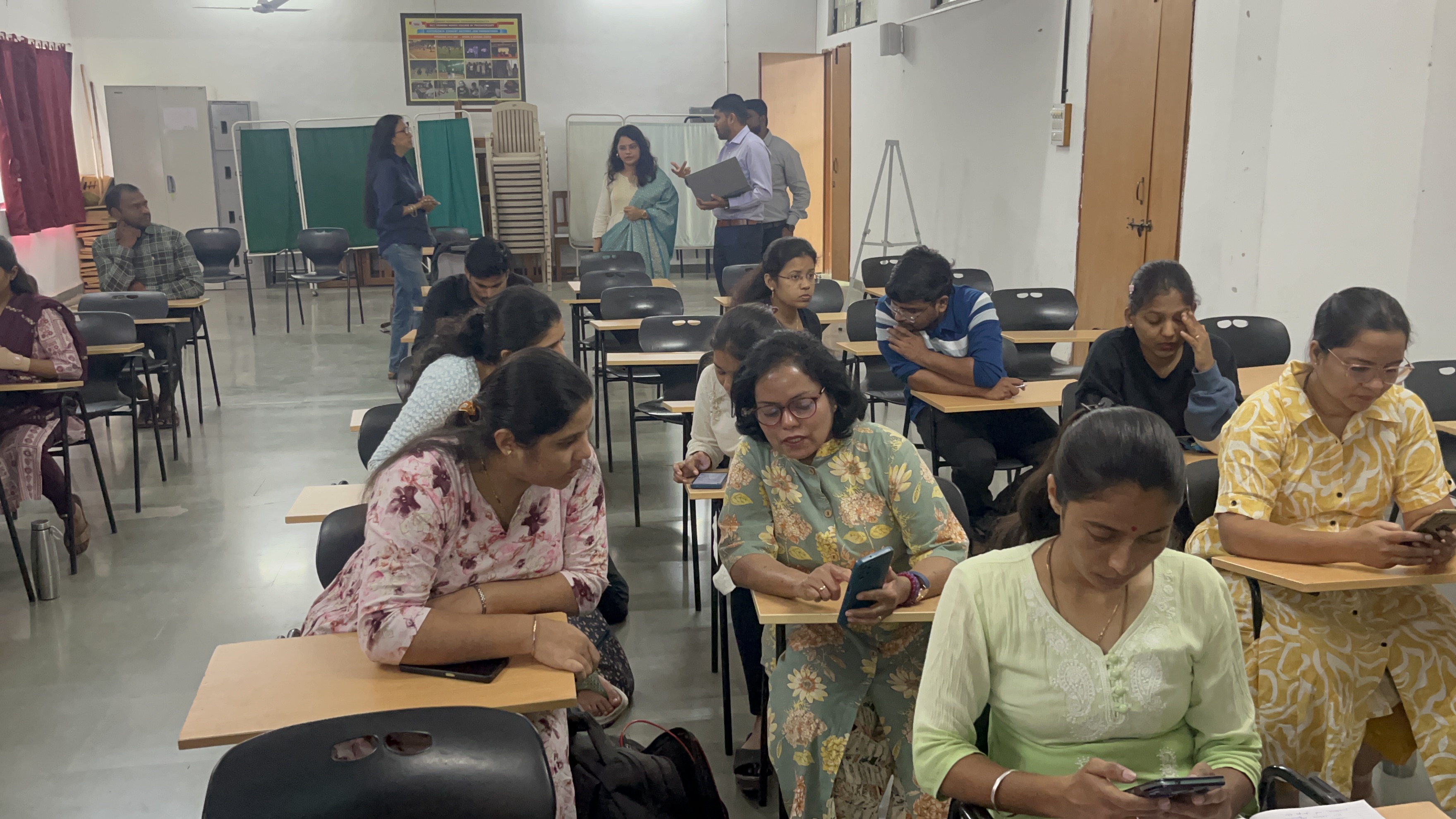 A group of people are seated at desks in a classroom or seminar setting, with some participants looking at their smartphones while others engage in discussion.