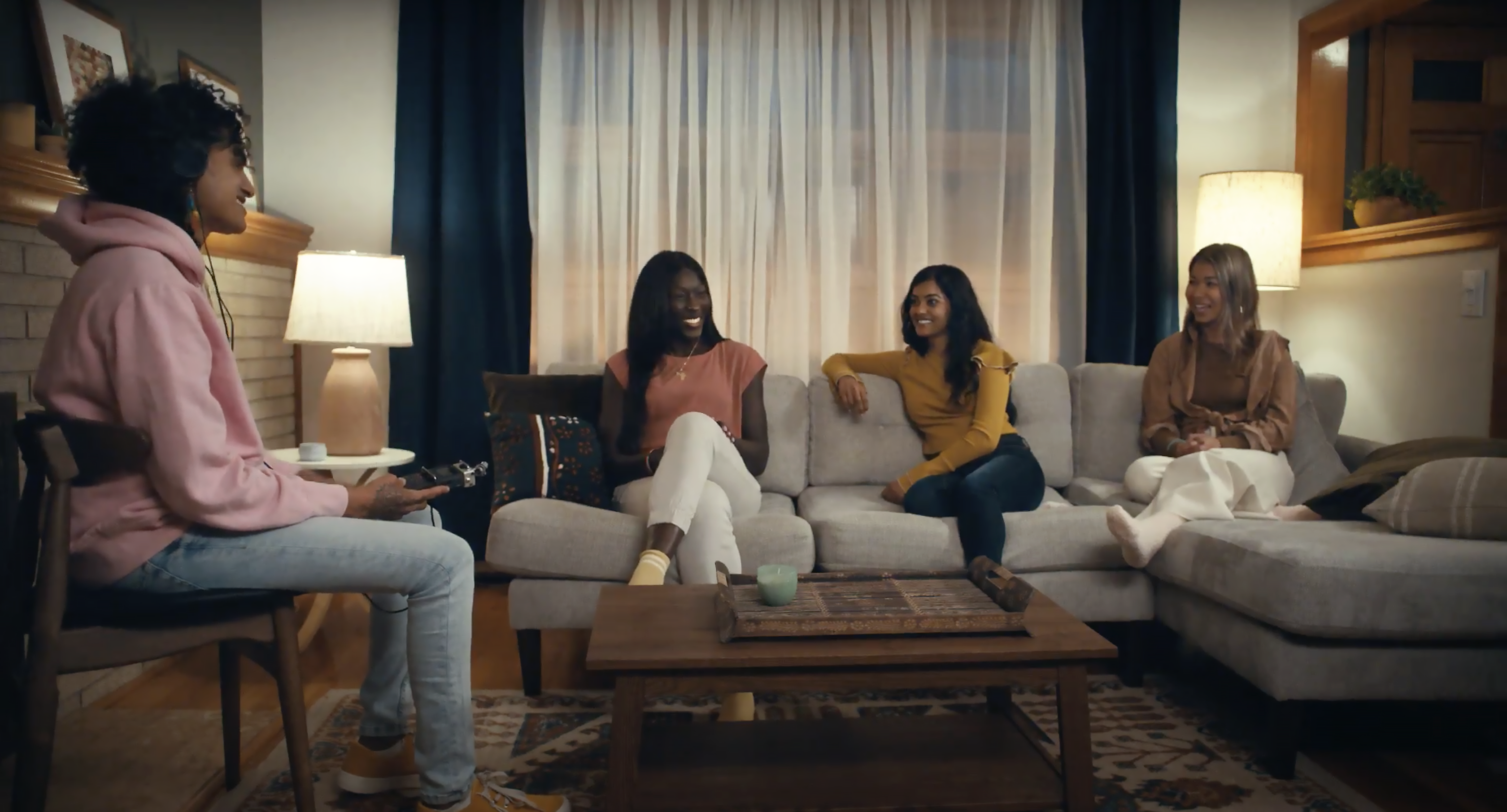 A person sits in a living room with a recording device. They're looking at and recording the conversation of three other people, people of color, who are sitting on a couch across the room from them.