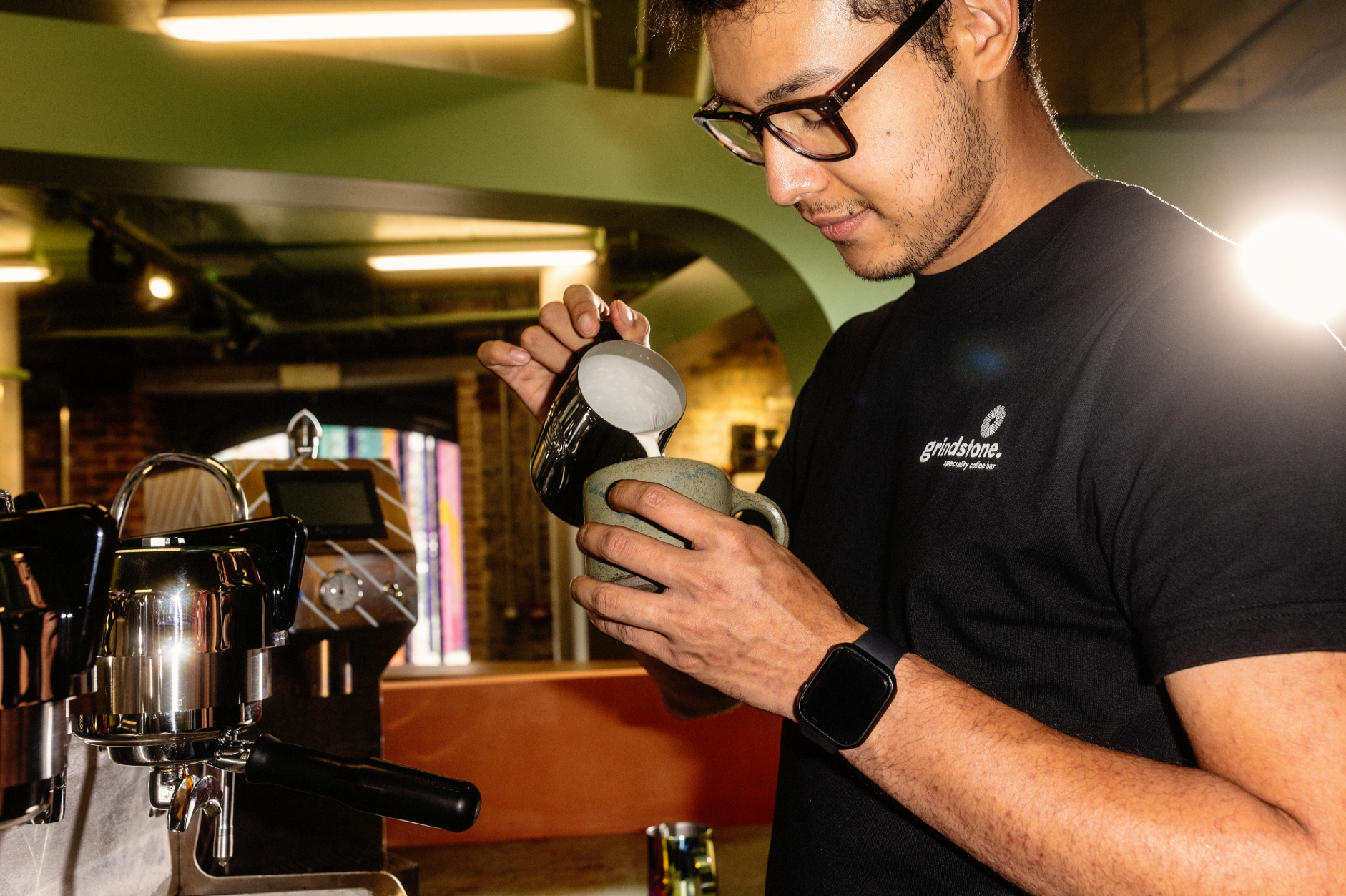 A barista at Grindstone Coffee carefully pouring steamed milk into a mug to make latte art.