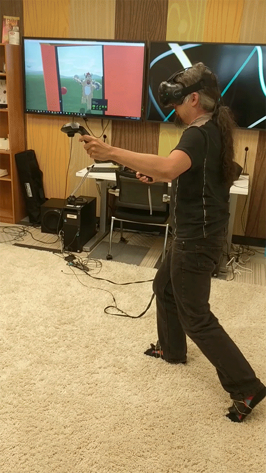 A live user with a VR headset and controllers moves his leg around the room. A monitor in the background reflects his in-game movement.