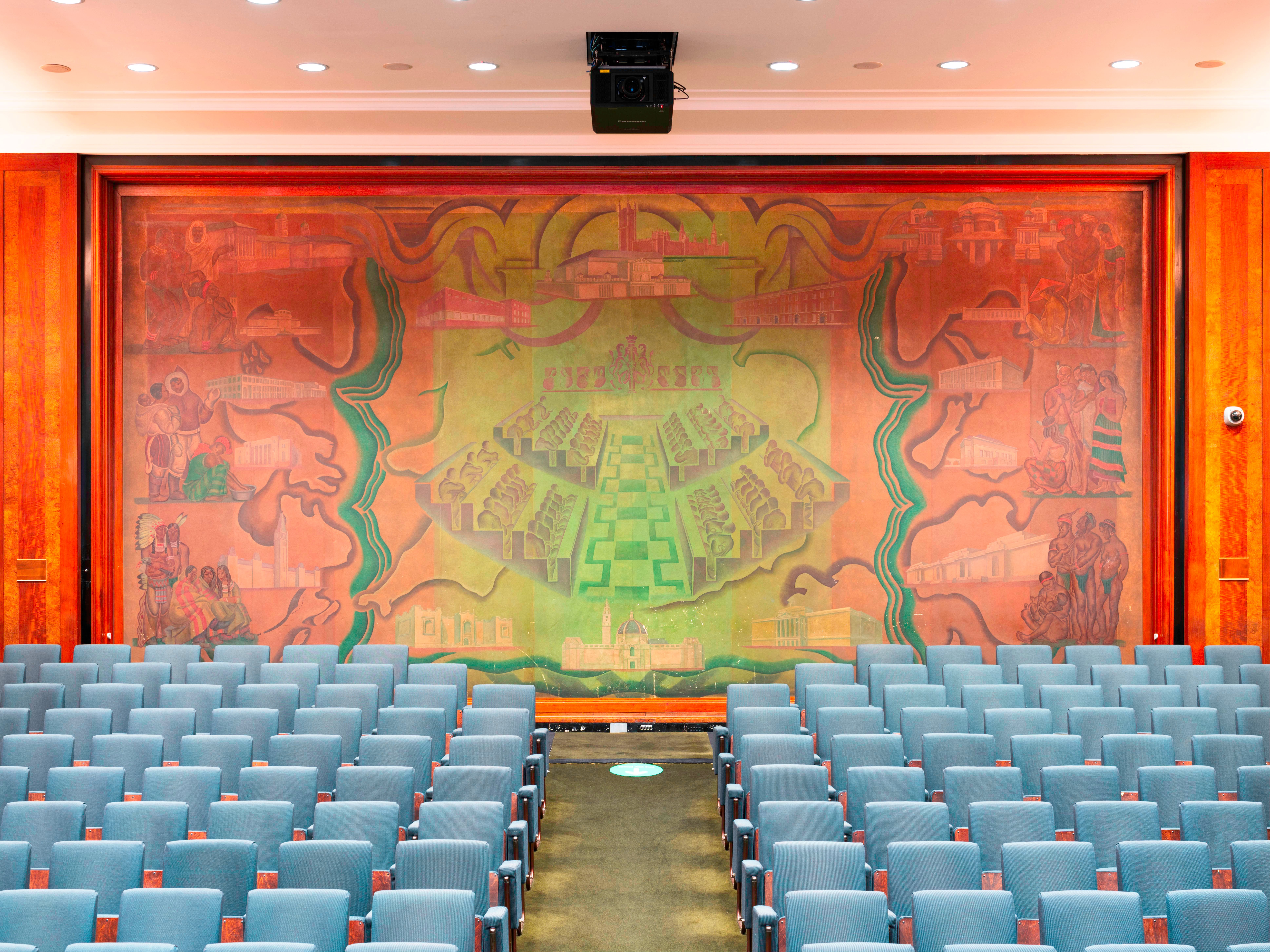 Large mural screen at the back of an auditorium with rows of empty blue seats.  The screen depicts a central hall with rows of seats amidst scenes of people and architecture.