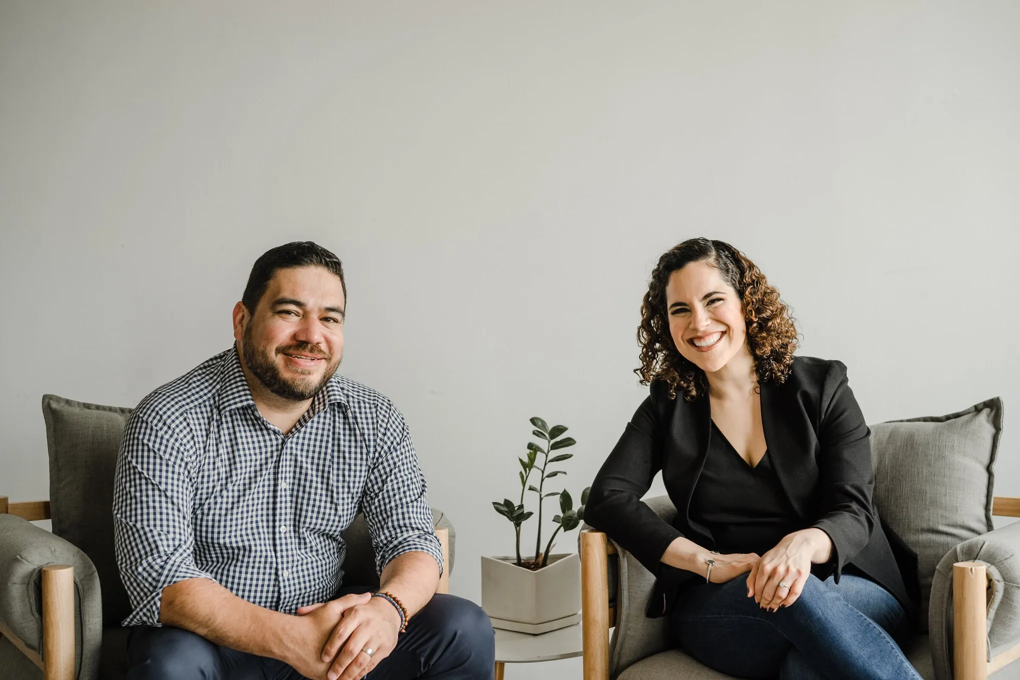 A man in a blue shirt and a woman in a black blazer sit side by side, hands folded,  in matching chairs in front of a gray wall.