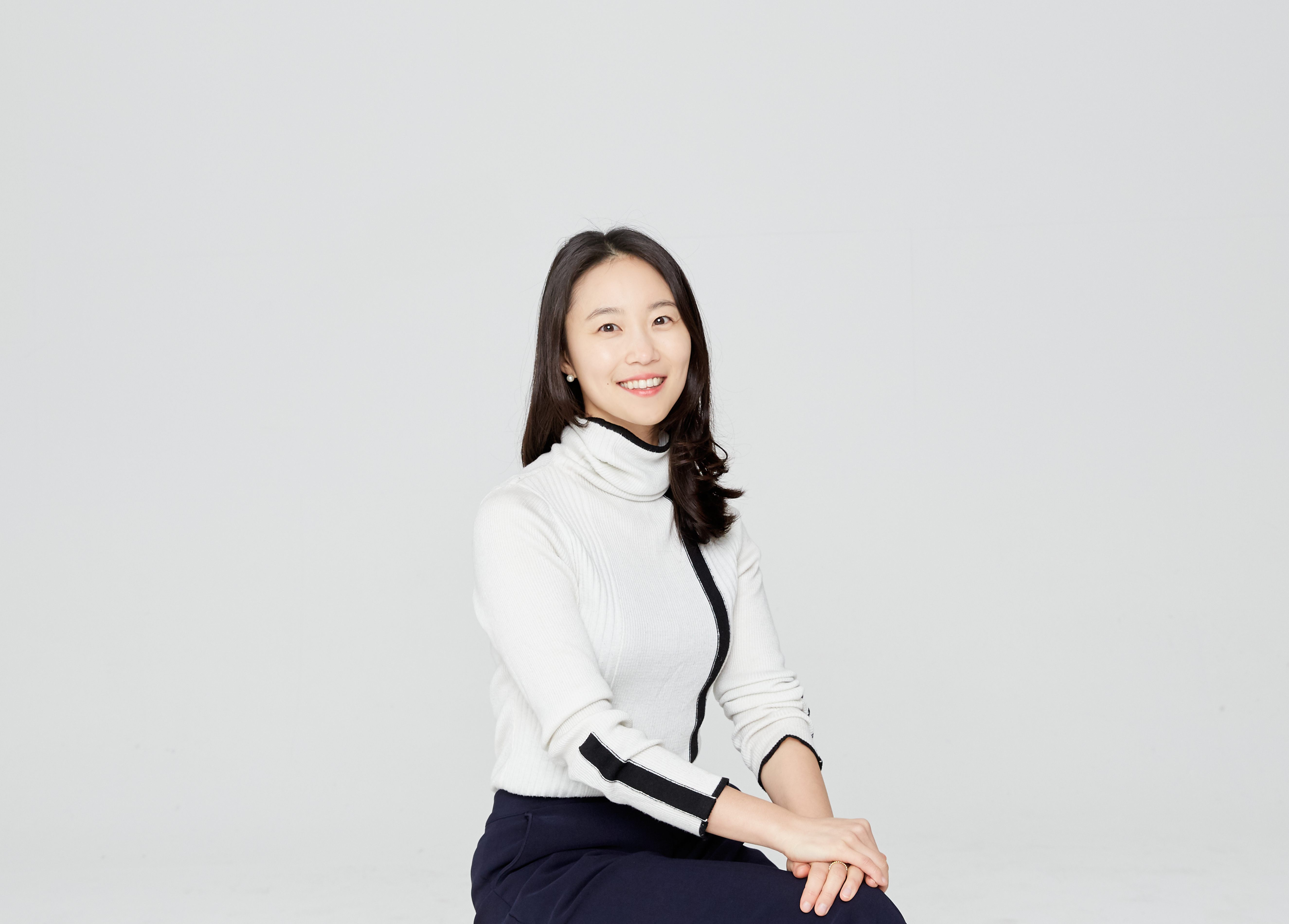 A woman wearing a white shirt and black trousers sits against a gray background with her hands crossed on her lap.