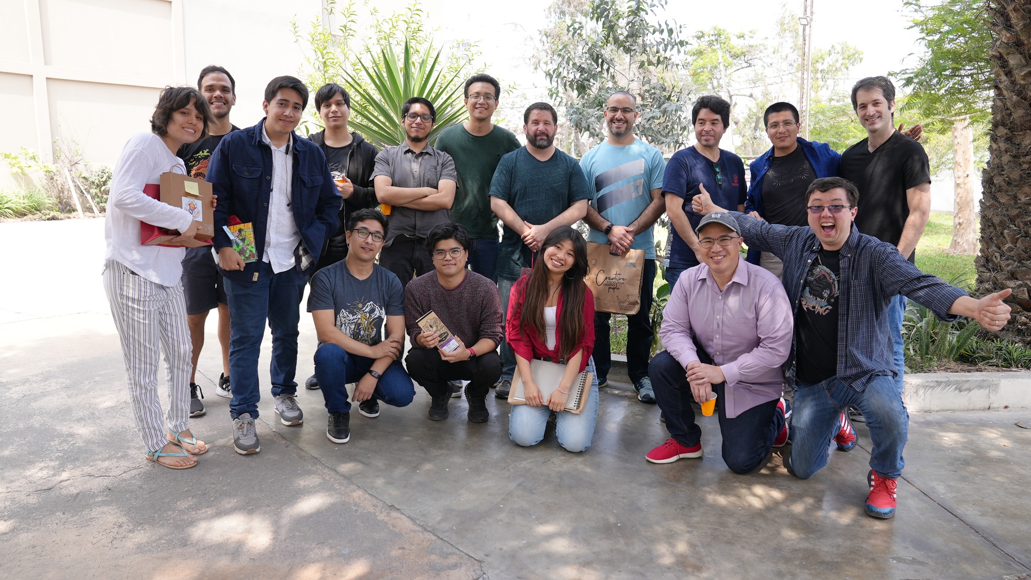 Sixteen members of Leap Game Studios pose for a picture on a bright day  in front of trees and foliage. The rightmost team member has their arms out wide with an excited expression.