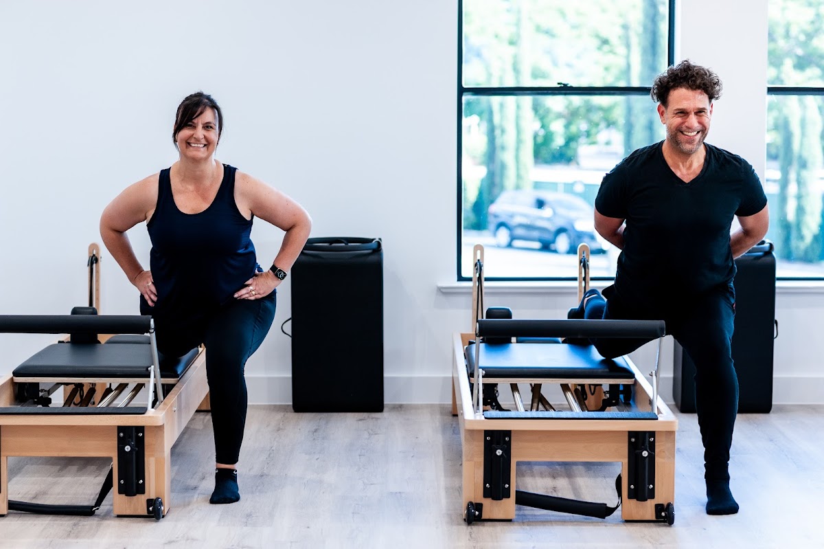 Two trainers are posing in front of reformer Pilates machines in a bright and modern fitness studio.